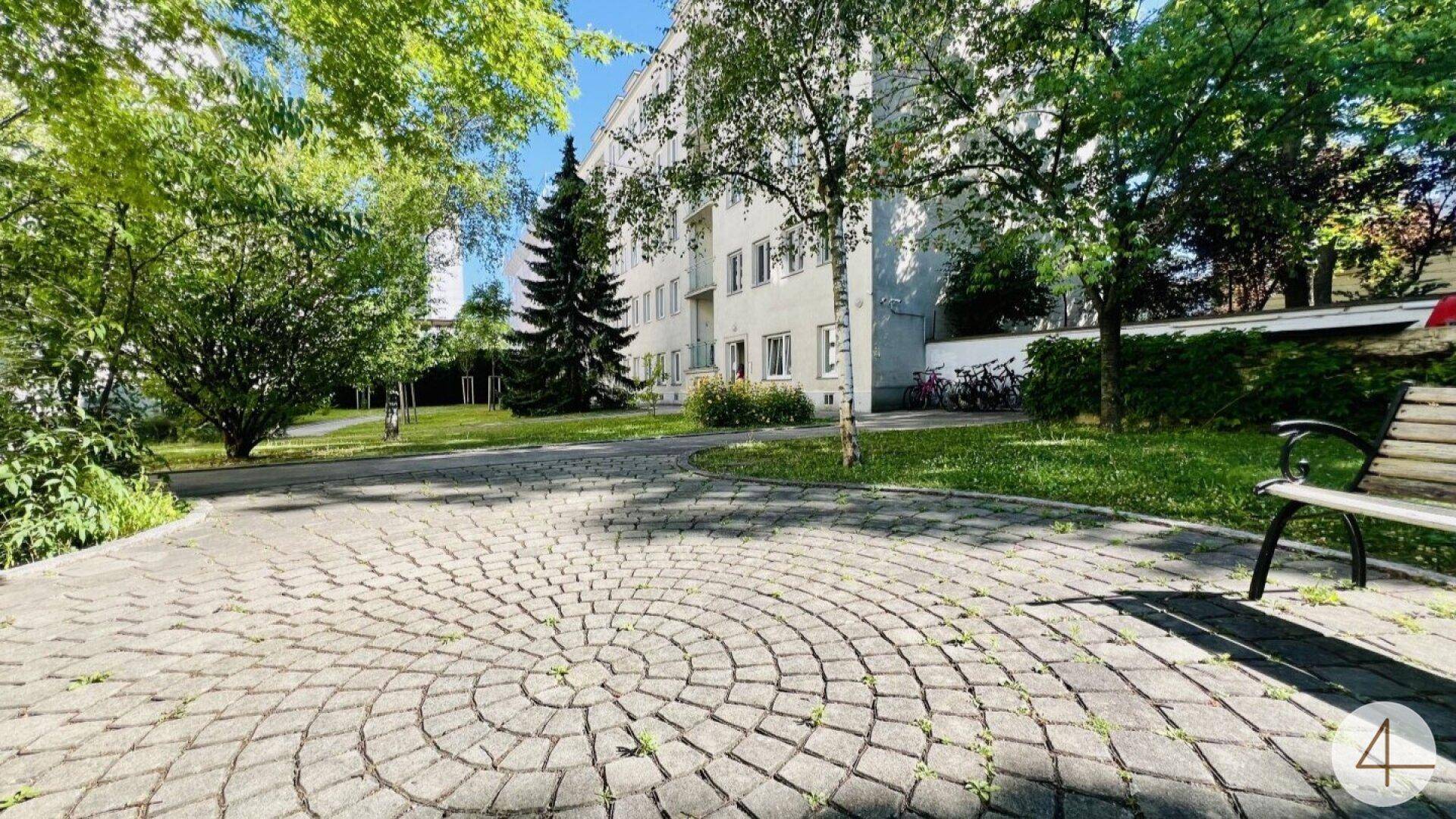 Gepflasterter Außenbereich mit Bäumen, Grünflächen und einem Mehrfamilienhaus im Hintergrund.