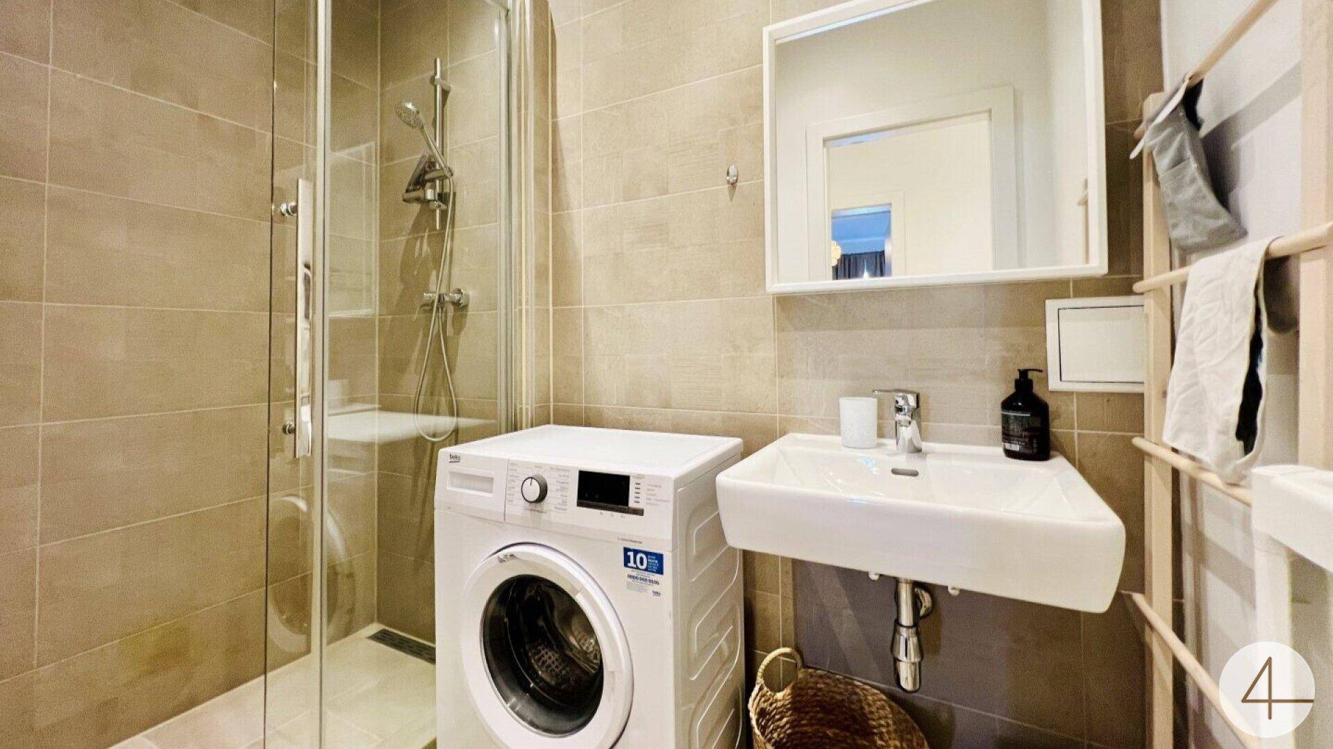 Modernes Badezimmer mit beigefarbenen Fliesen, begehbarer Dusche und Waschmaschine.