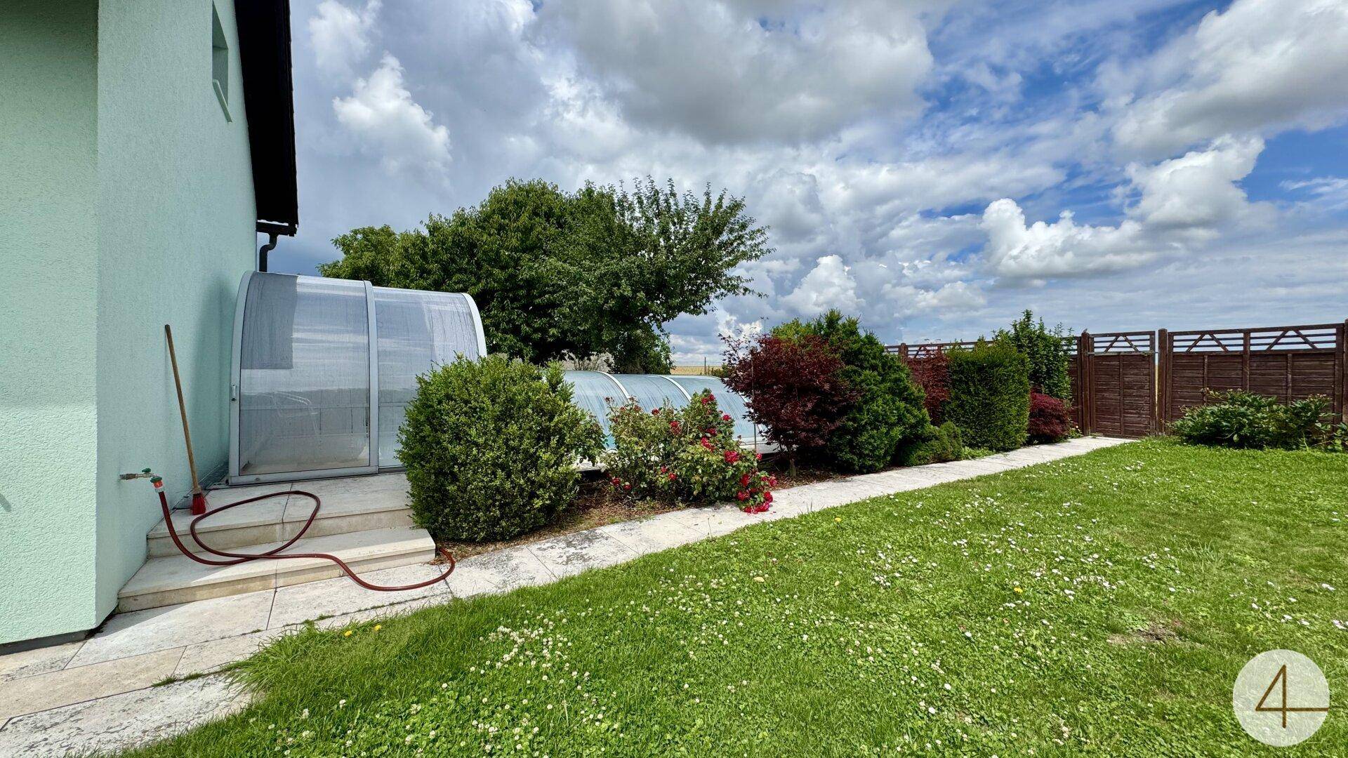 Seitenansicht des Hauses mit Rasenfläche, Gartenbepflanzung und dem überdachten Poolbereich im Hintergrund.