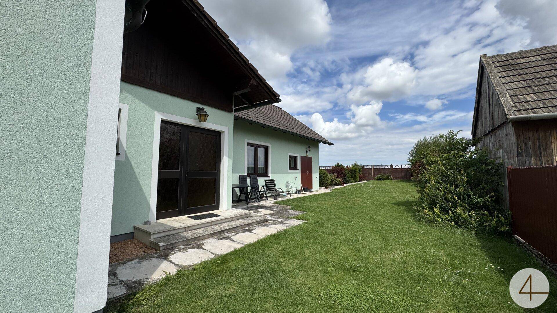 Außenansicht des Hauses mit einer kleinen Terrasse, Rasenfläche und Gartenmöbeln unter bewölktem Himmel.