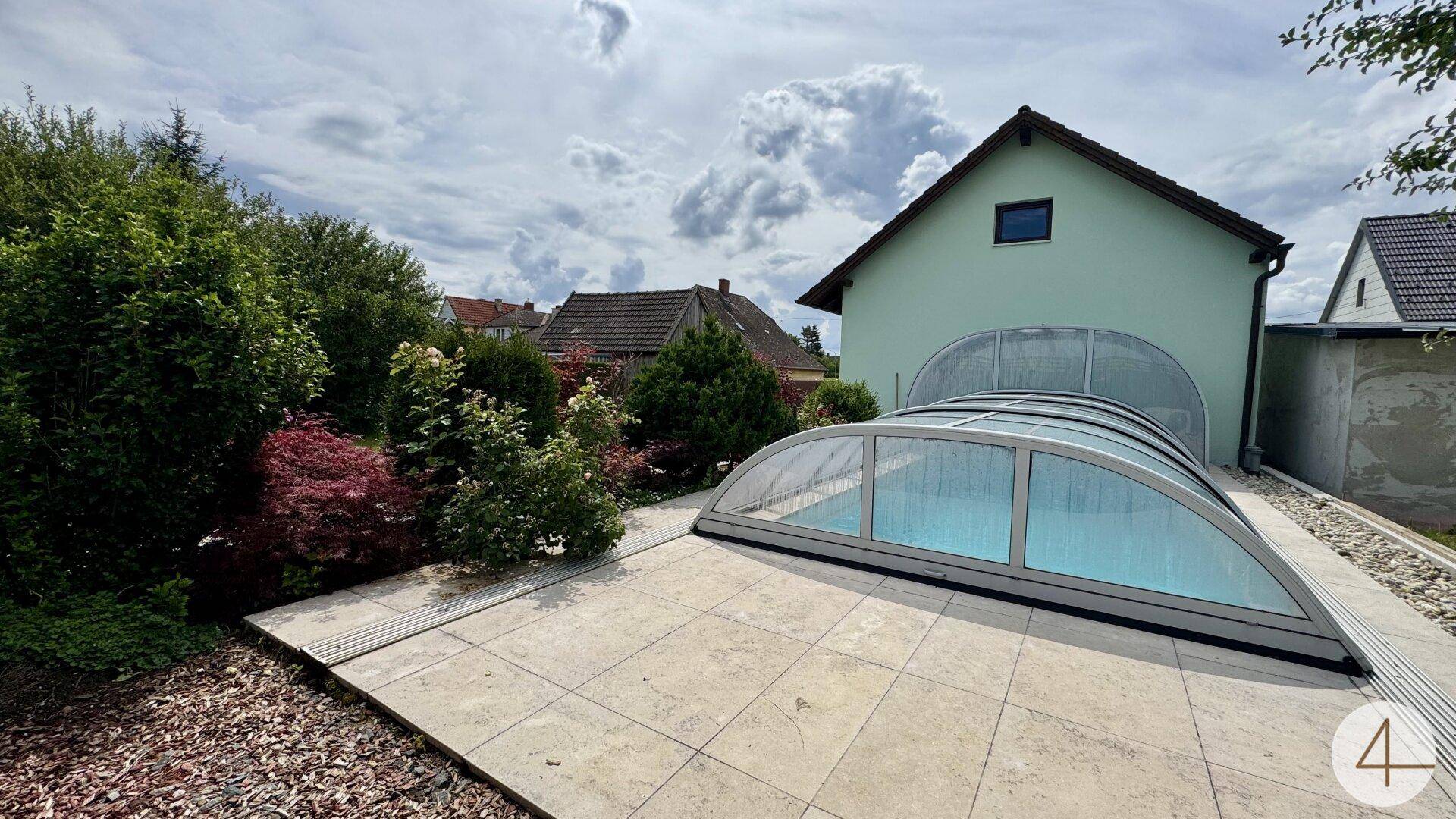 Blick auf das Haus und den überdachten Poolbereich mit gefliester Terrasse und umgebender Bepflanzung.
