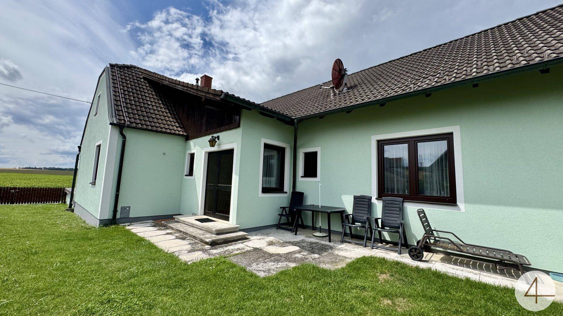 Außenbereich des Hauses mit möblierter Terrasse, Rasenfläche und Liegestuhl unter bewölktem Himmel.