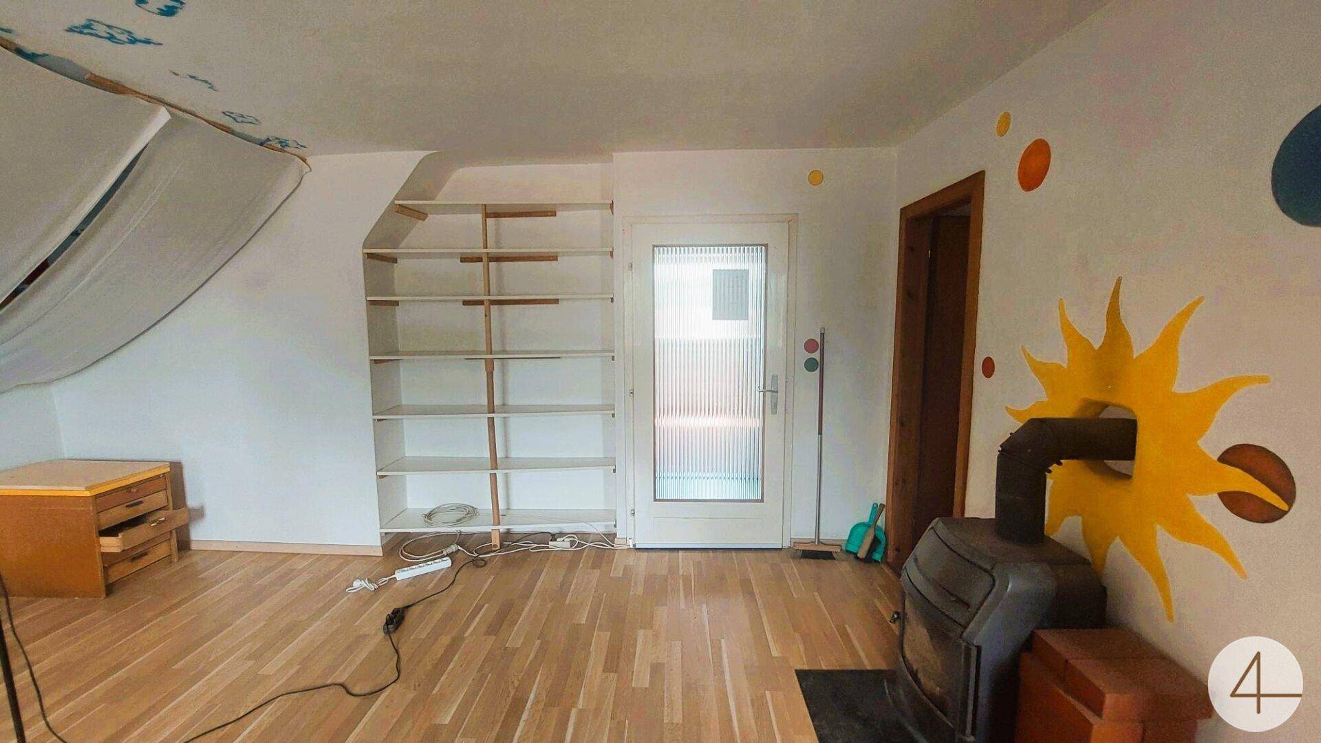 Heller Wohnbereich mit Kaminofen, Einbauregalen und Zugang zu einem weiteren Raum.