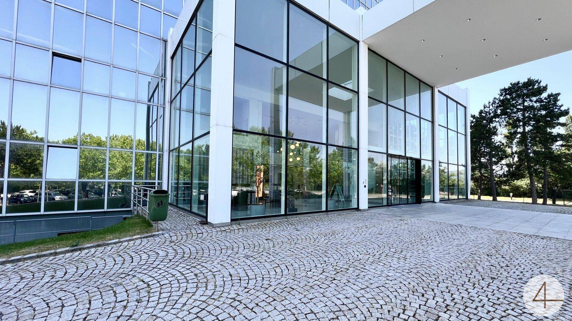 Eingangsbereich des modernen Bürogebäudes mit Glasfassade und gepflastertem Vorplatz.