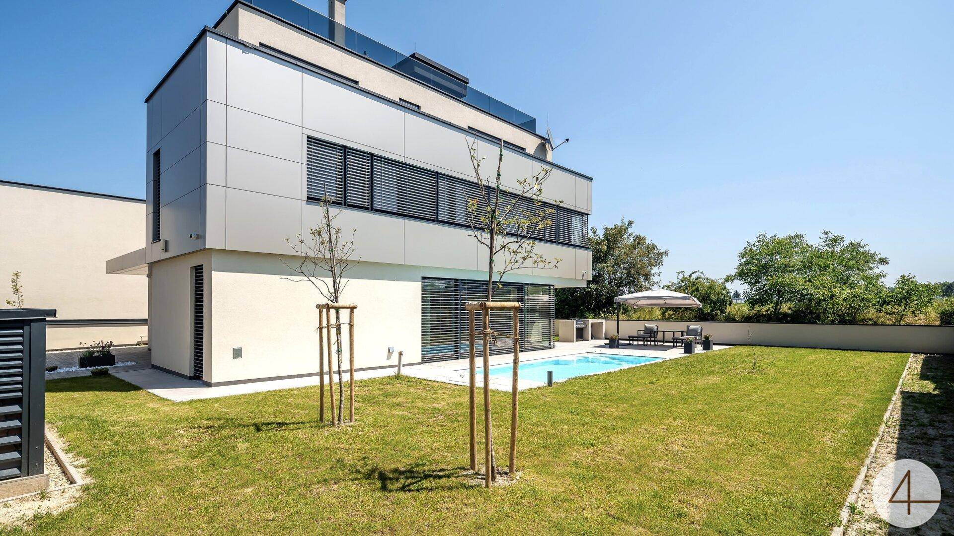 Seitliche Ansicht des modernen Hauses mit Poolbereich und weitläufigem Rasen unter blauem Himmel.