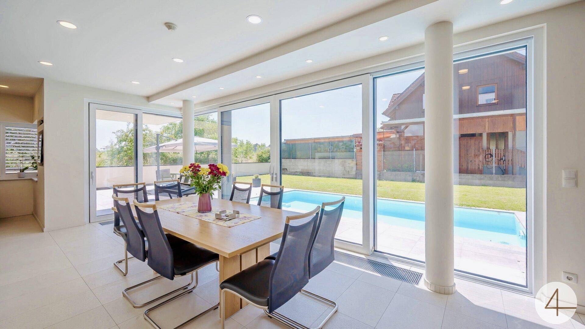 Heller Essbereich mit großem Holztisch und bodentiefen Fenstern mit Blick auf den Pool und Garten.
