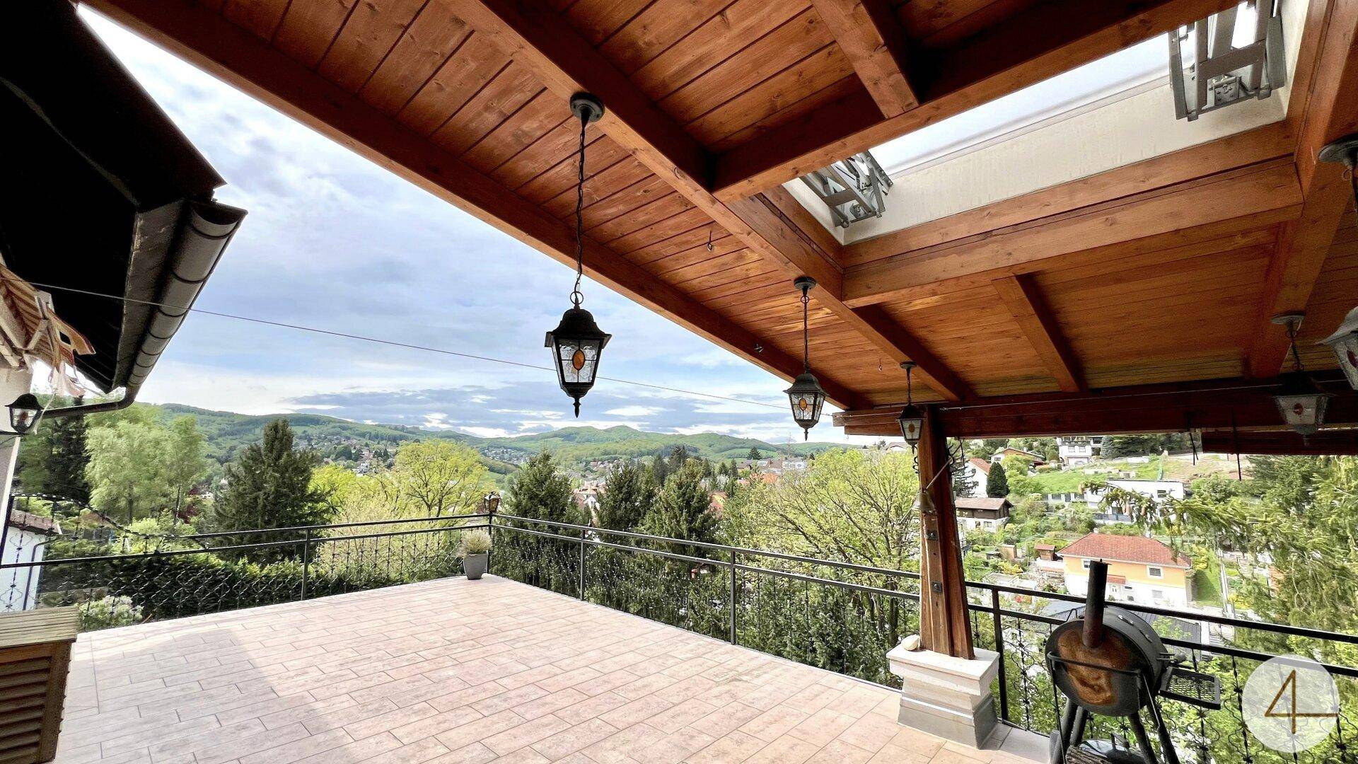 Überdachte Terrasse mit Holzbalken, Außenleuchten und weitem Blick über die grüne Umgebung.