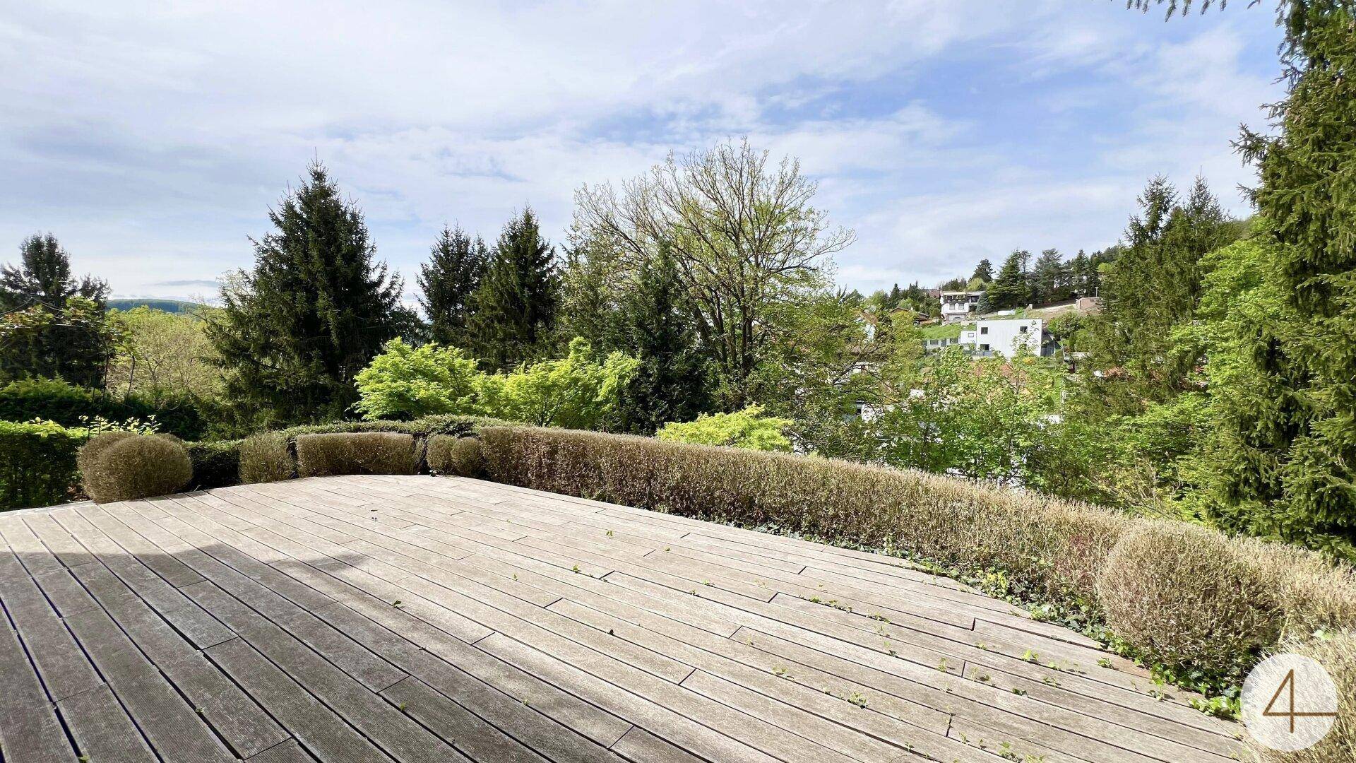 Große Holzterrasse umgeben von Hecken und Bäumen mit Blick ins Grüne.
