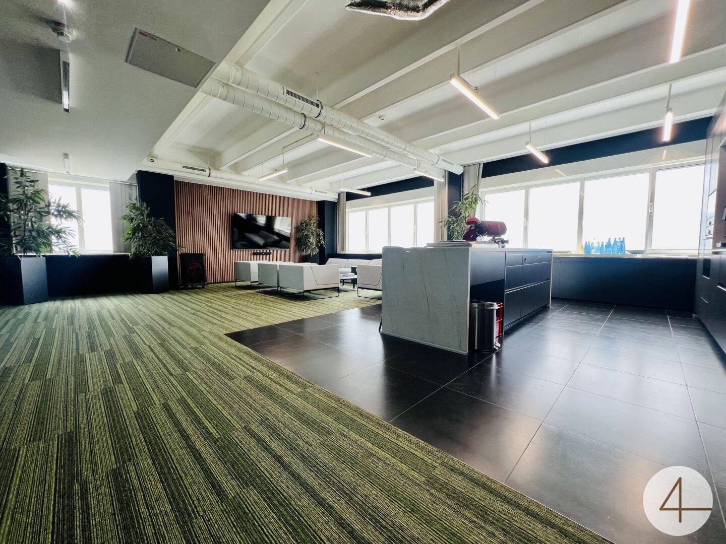 Offener Bürobereich mit Lounge-Sitzgelegenheiten, einer modernen Küchenzeile und großen Fenstern.