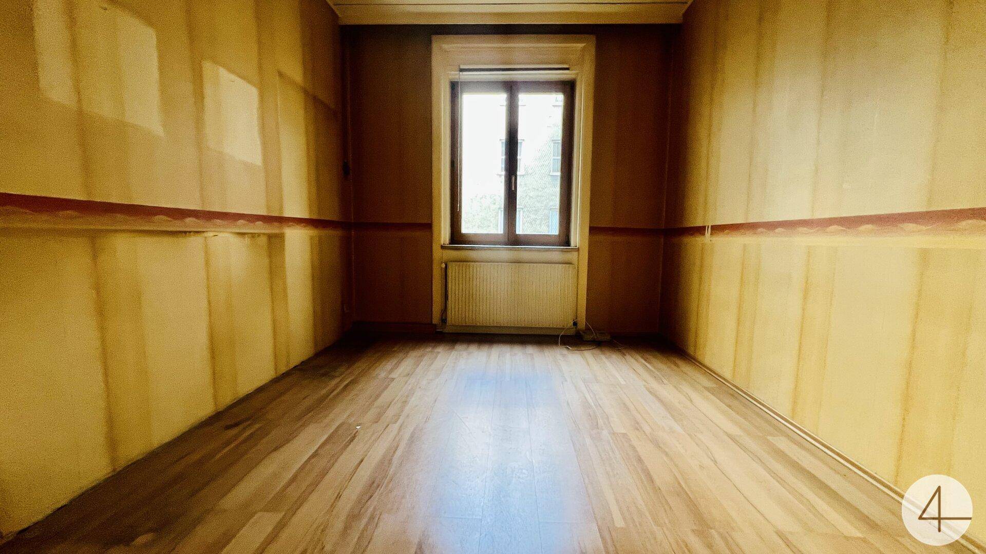 Leeres Zimmer mit Laminatboden und renovierungsbedürftigen Wänden, hell durch ein Fenster.
