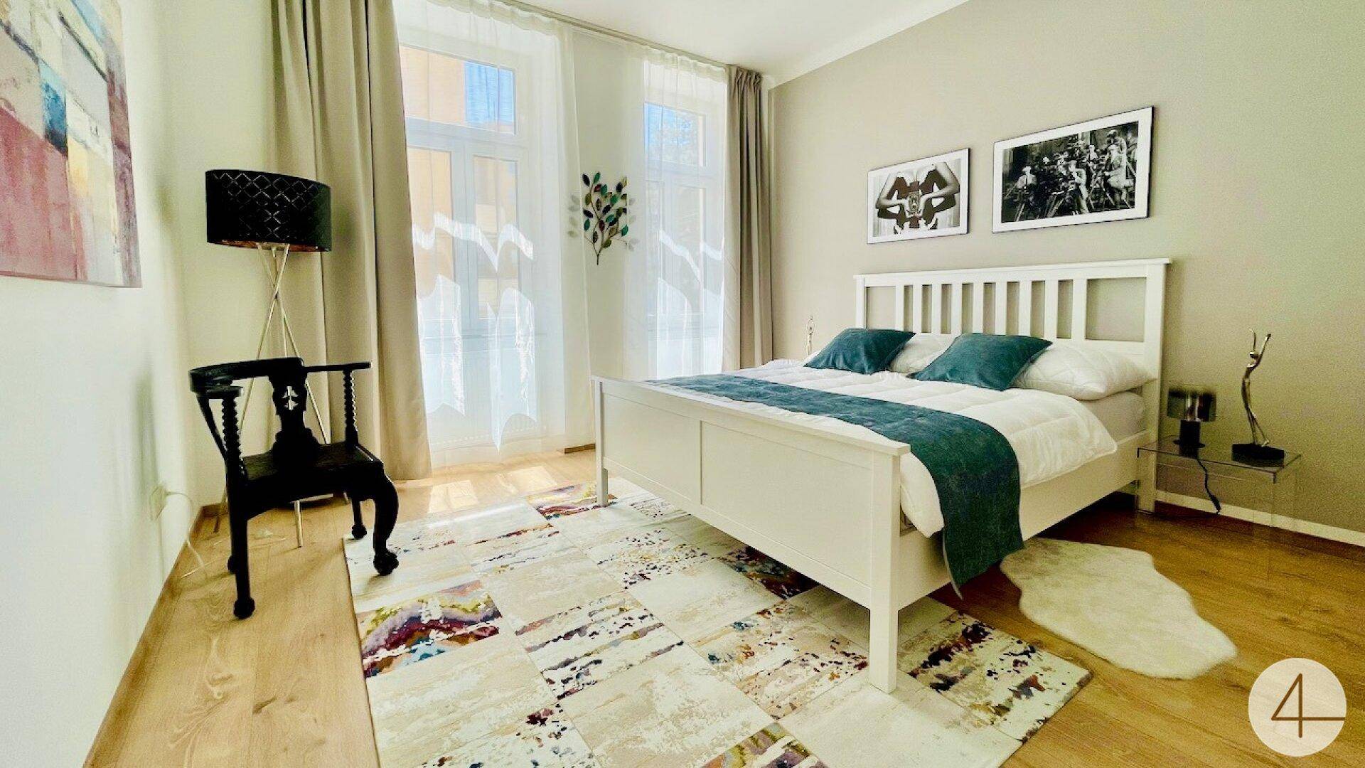 Gemütliches Schlafzimmer mit weißem Bett, Holzboden und großem Fenster für viel Tageslicht.
