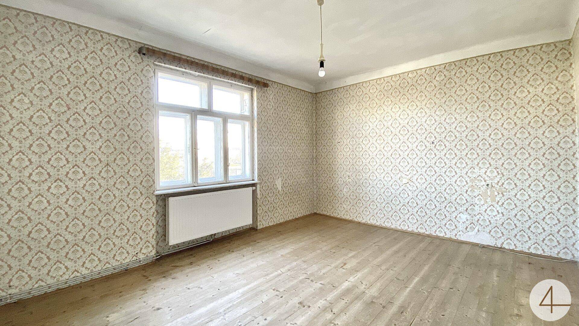 Schlafzimmer mit gemusterter Tapete, Holzdielenboden und einem großen Fenster für Tageslicht.
