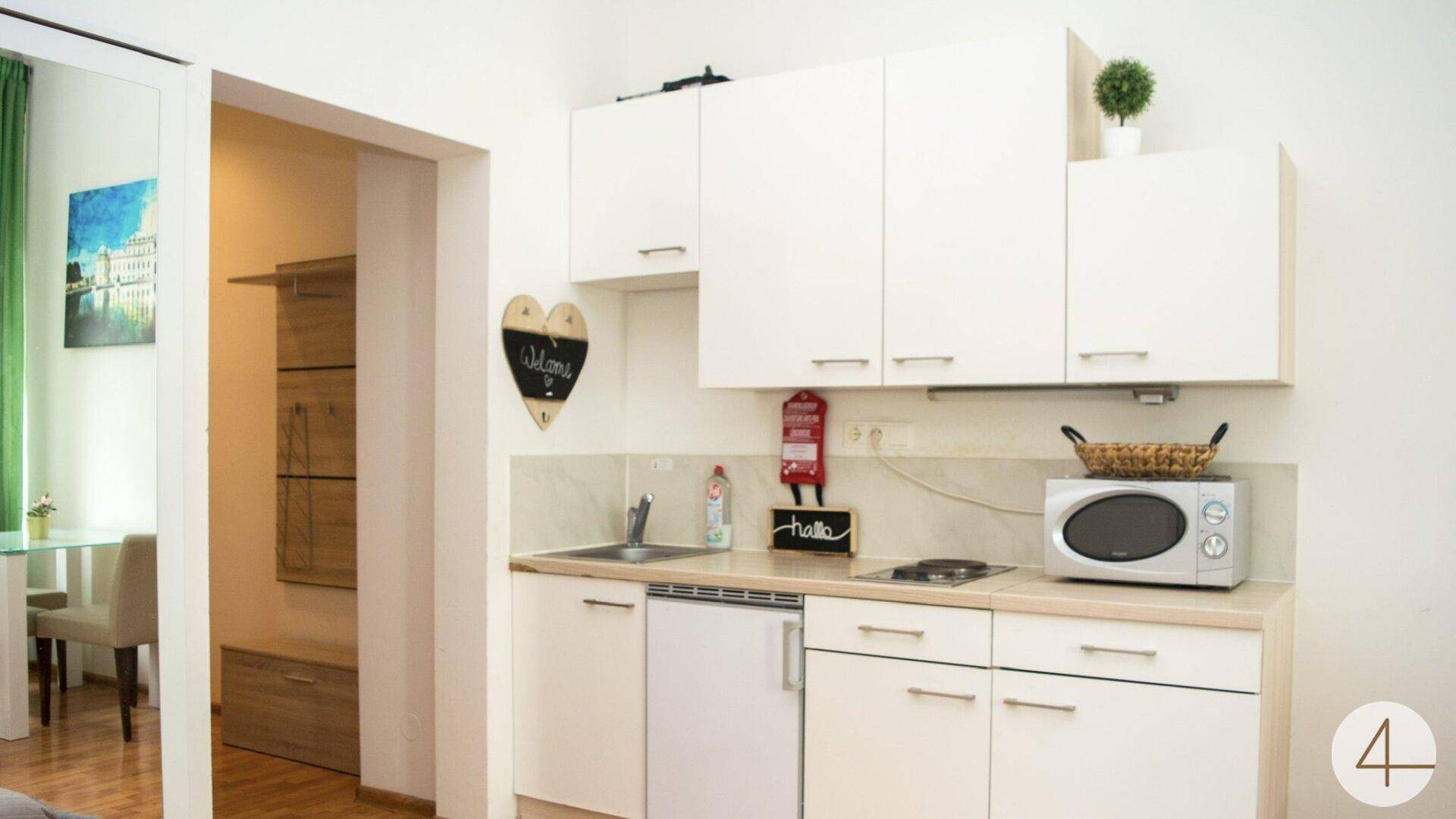 Kleine weiße Küchenzeile mit Mikrowelle, Kochfeld und Hängeschränken in einem Studioapartment.