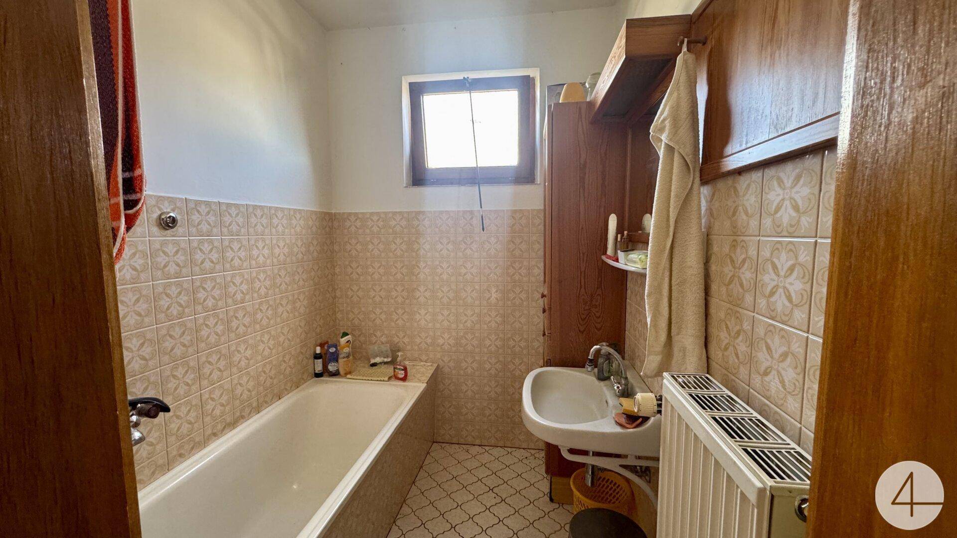 Badezimmer mit Badewanne, Waschbecken, Fenster und hellbraunen Fliesen im traditionellen Stil.