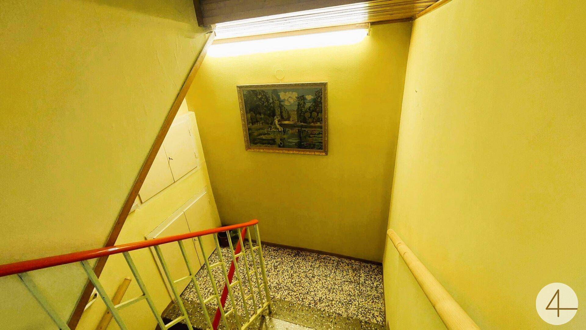 Treppenhaus mit gelben Wänden, Terrazzoboden und einfacher Beleuchtung.