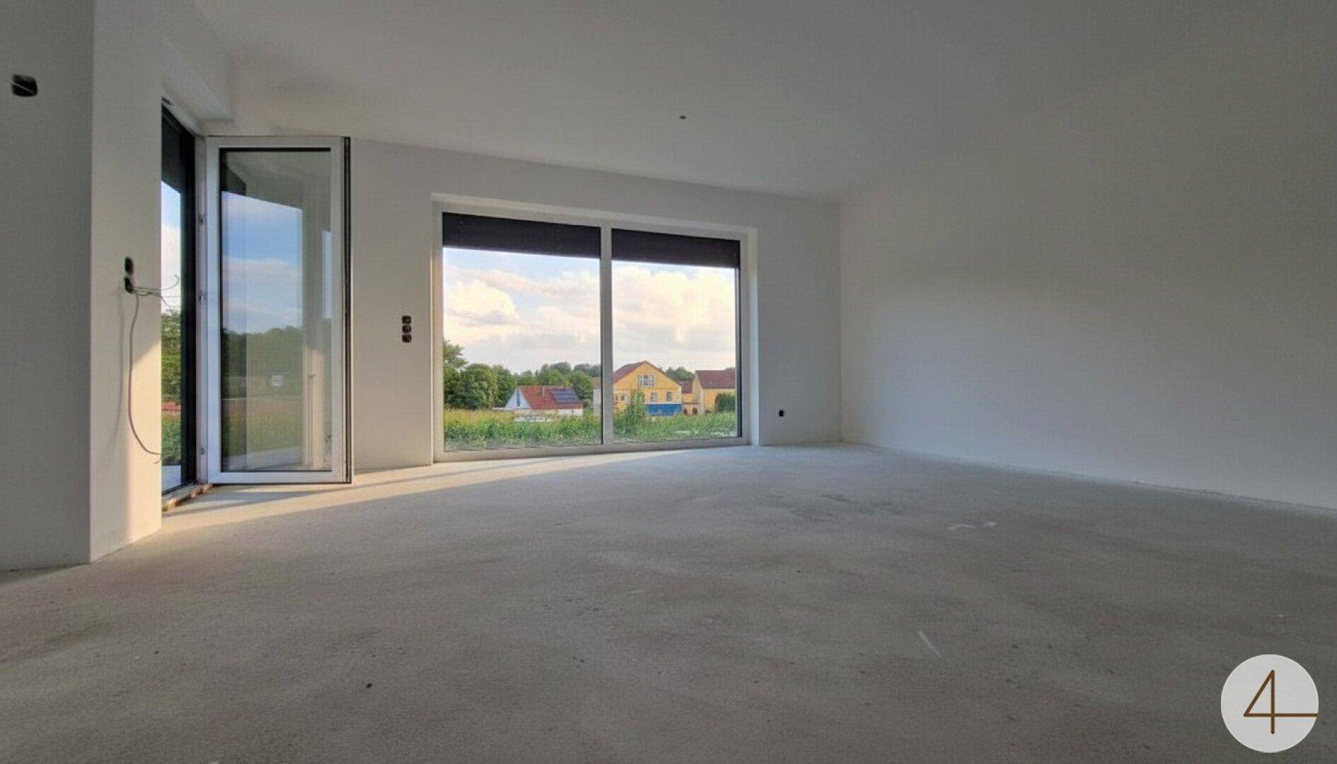 Großer, heller Raum im Rohbau mit Schiebetüren und Blick auf die umliegende Landschaft und Nachbarhäuser.