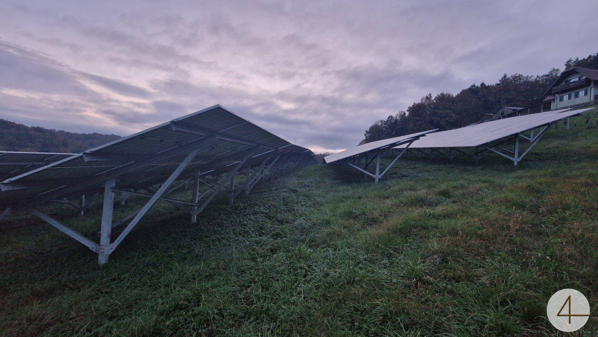 Solarmodule auf einem begrünten Hang unter bewölktem Himmel, mit einem Haus im Hintergrund.