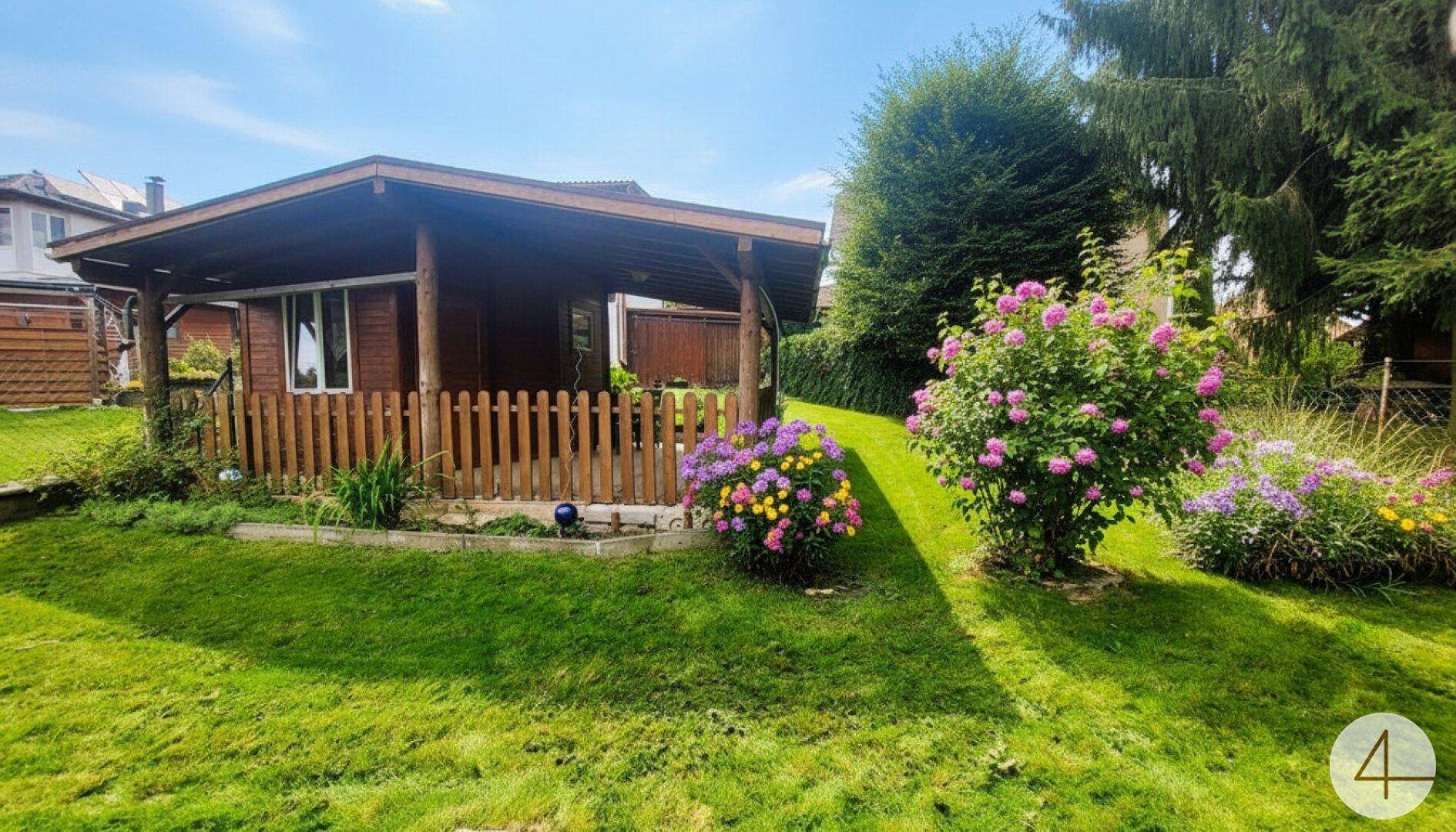 Gemütliches Gartenhaus aus Holz mit überdachter Terrasse, umgeben von blühenden Sträuchern und Rasen.
