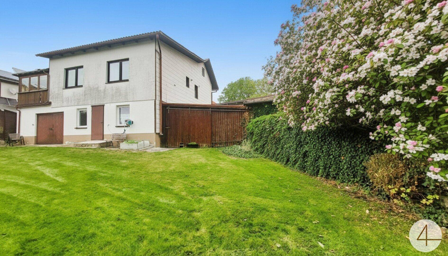 Großer grüner Garten mit Rasenfläche und blühenden Bäumen hinter dem weißen Wohnhaus.