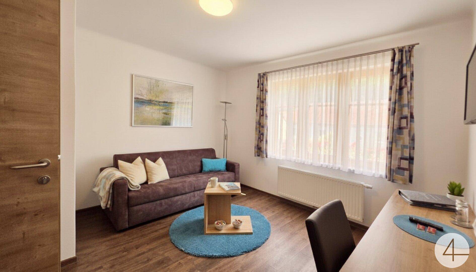 Gemütliches Wohnzimmer mit Sofa, Schreibtisch und großem Fenster für viel Tageslicht.