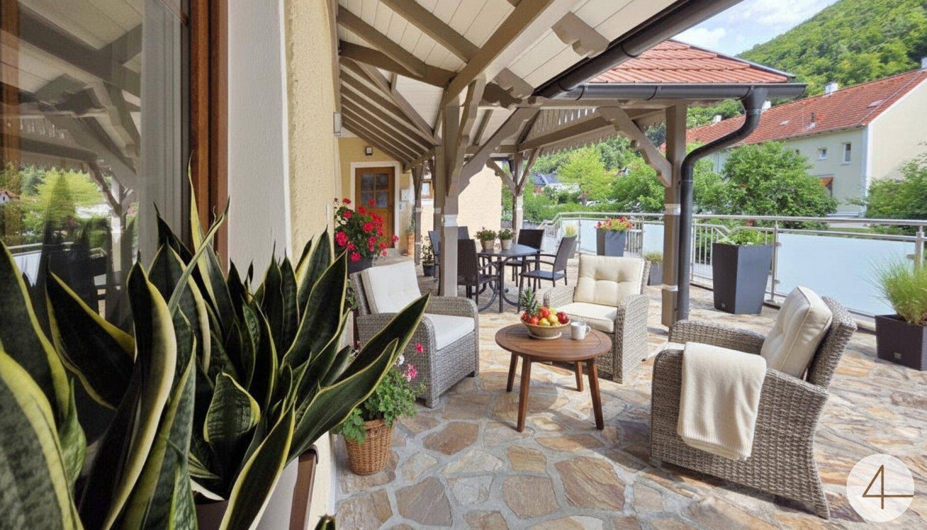 Überdachte Terrasse mit bequemen Rattanmöbeln, Steinboden und Blick ins Grüne.