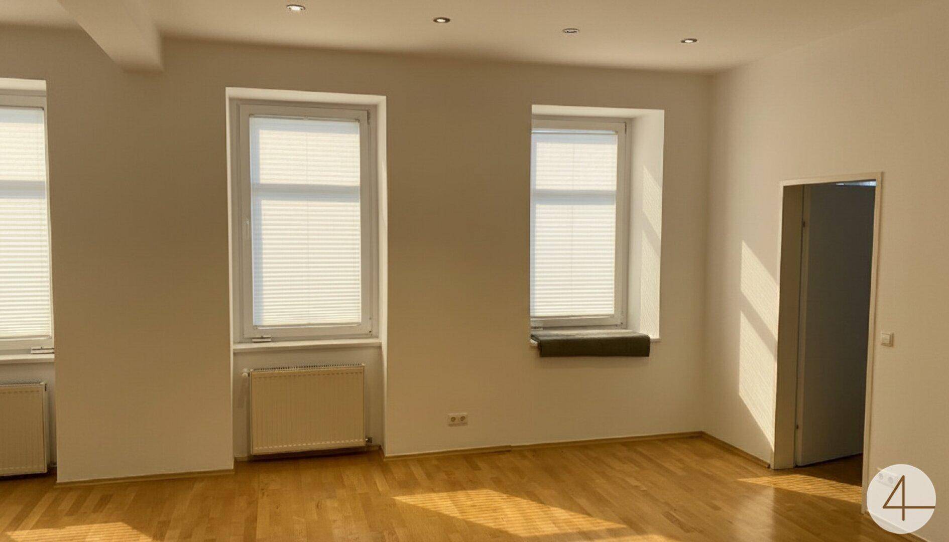 Großer, heller Raum mit Parkettboden, mehreren Fenstern mit Jalousien und viel Tageslicht.