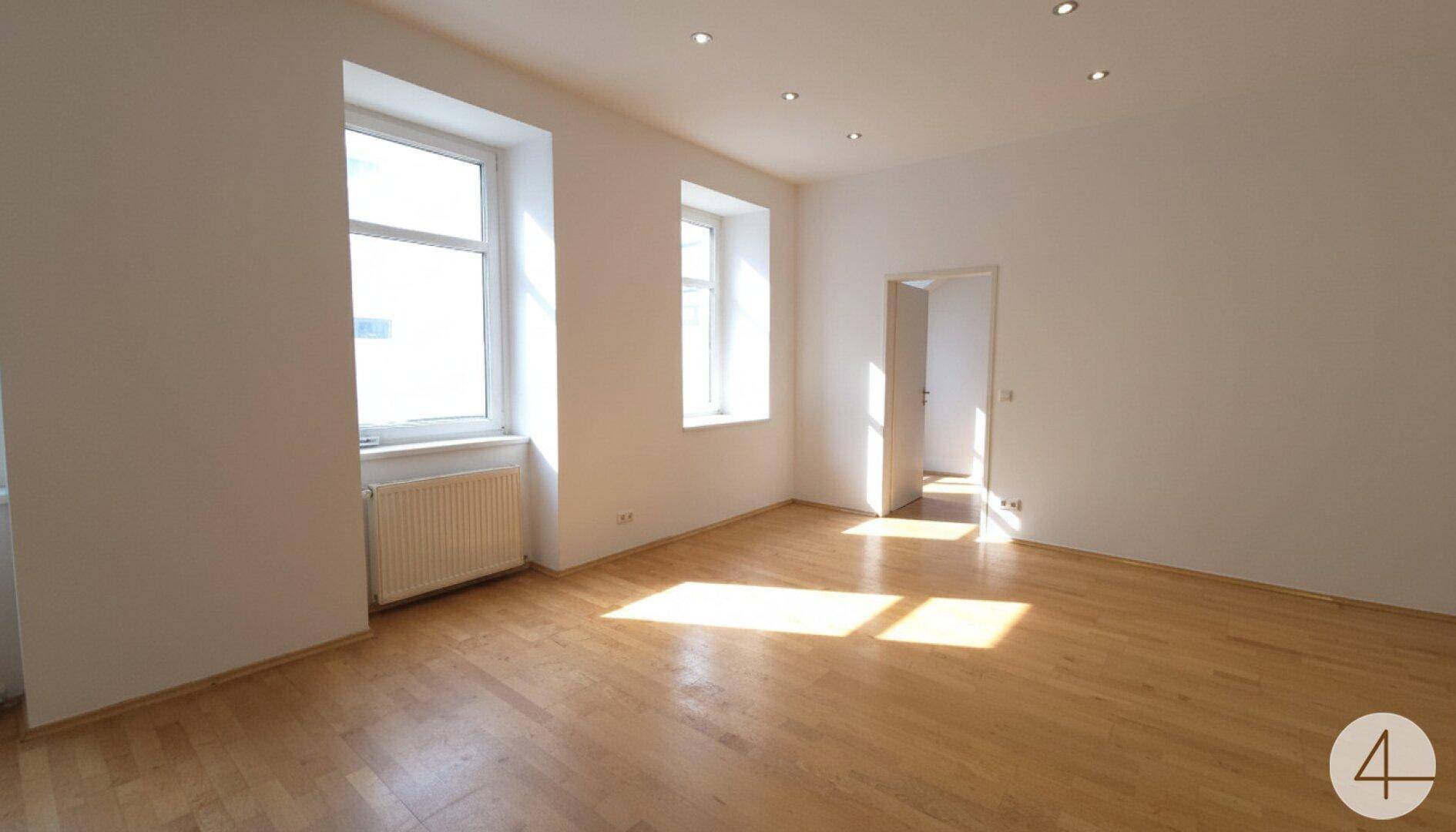 Geräumiges Wohnzimmer mit hellem Parkettboden, großen Fenstern und viel natürlichem Tageslicht.
