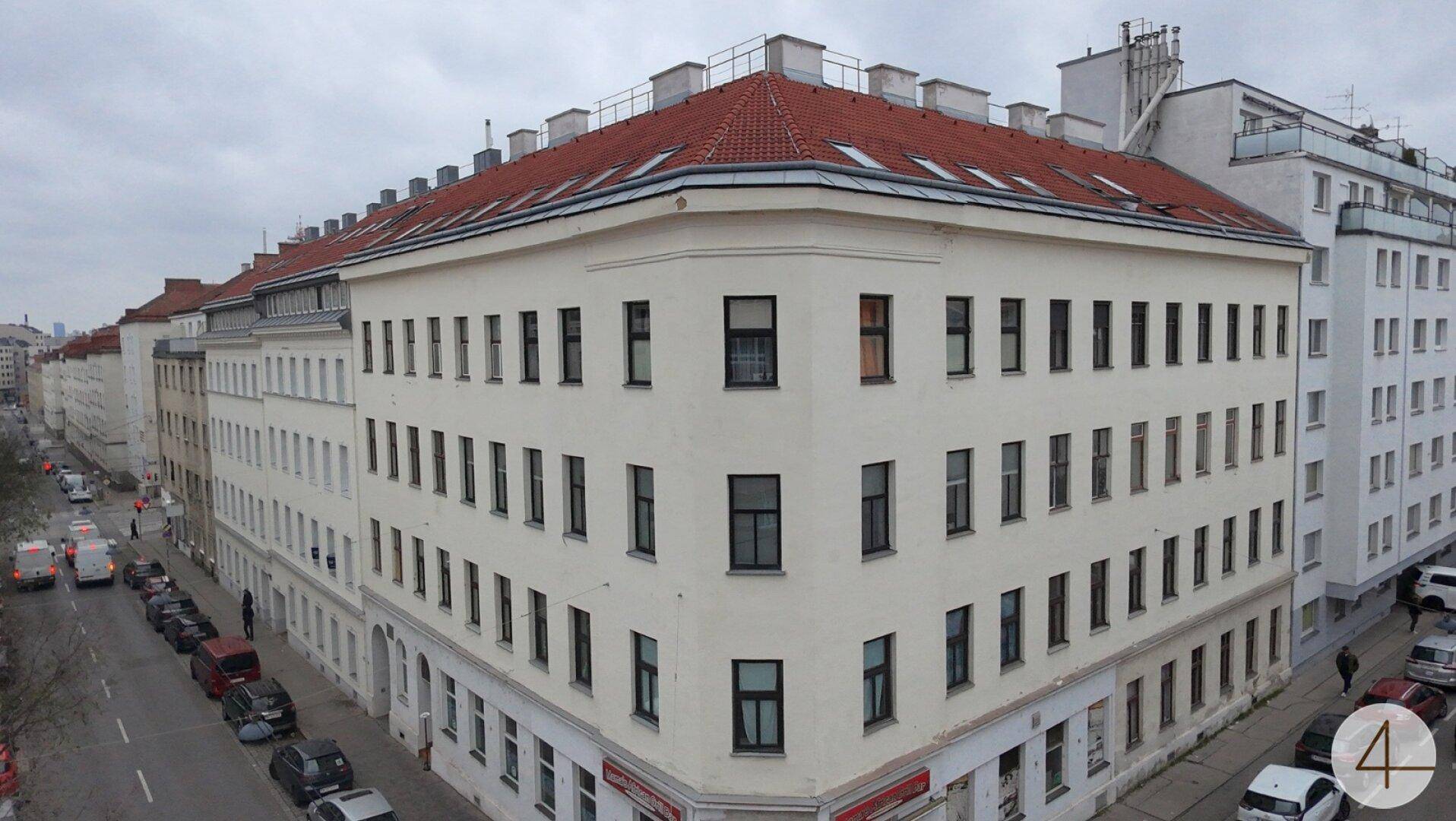Historisches Eckgebäude mit klassischer Architektur und rotem Ziegeldach in städtischer Umgebung.