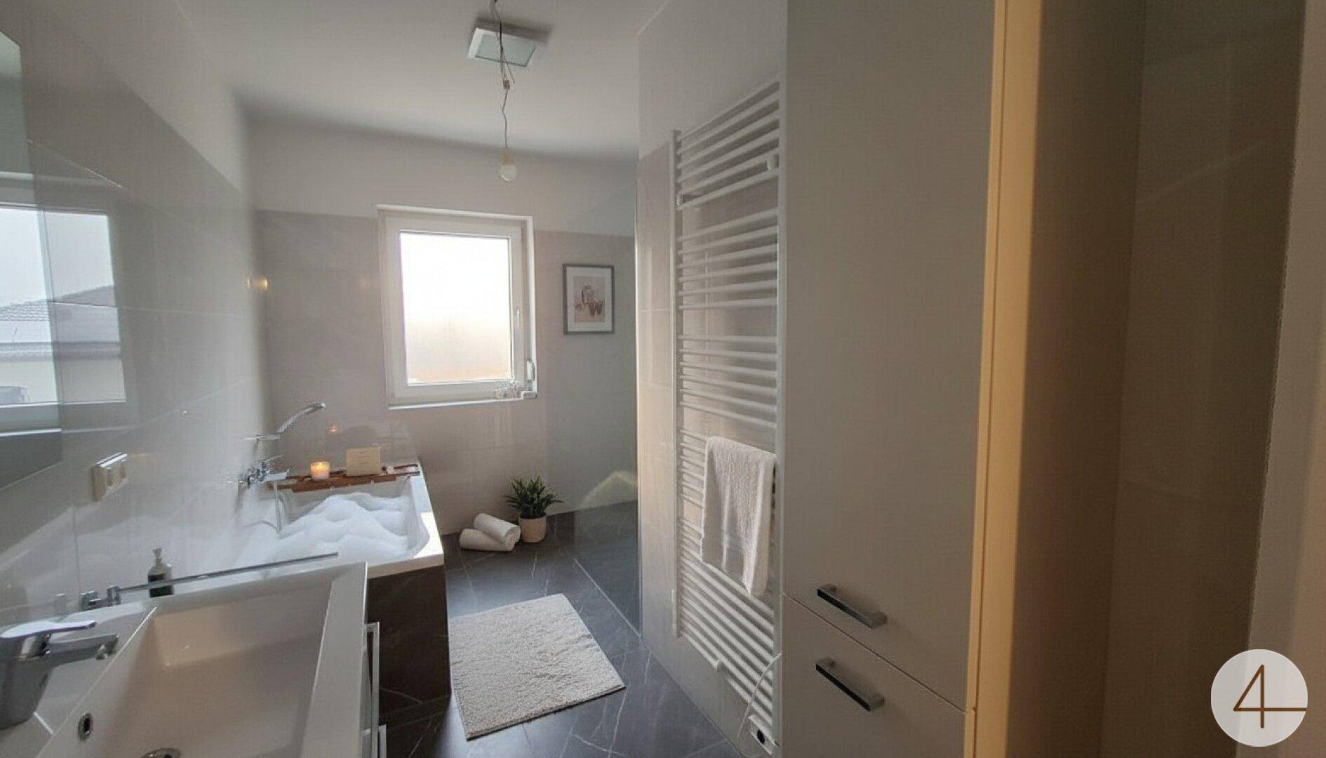 Modernes Badezimmer mit Badewanne, Fenster und beheiztem Handtuchhalter in hellgrauen Fliesen.