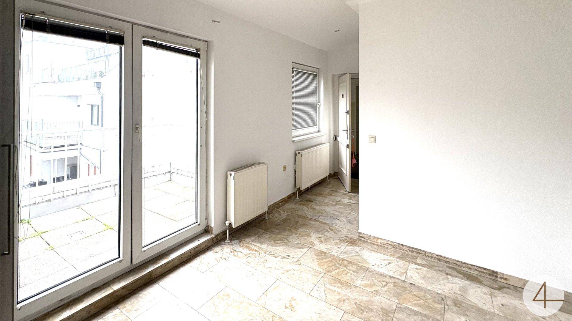 Heller Raum mit Fliesenboden und großen Fenstertüren, die zu einem Außenbereich führen.