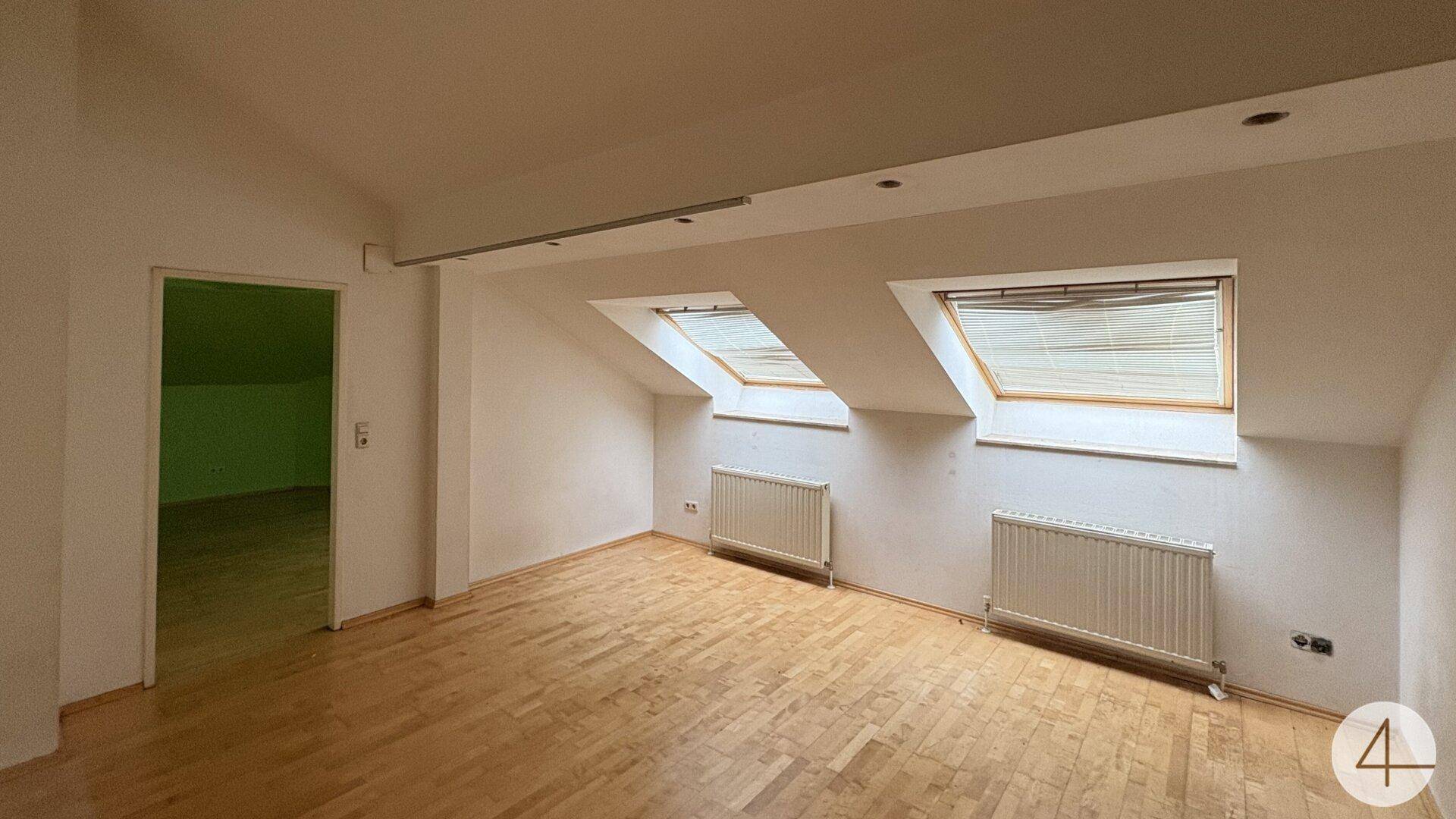 Helles Dachzimmer mit schrägen Wänden, Dachfenstern und Holzparkettboden.