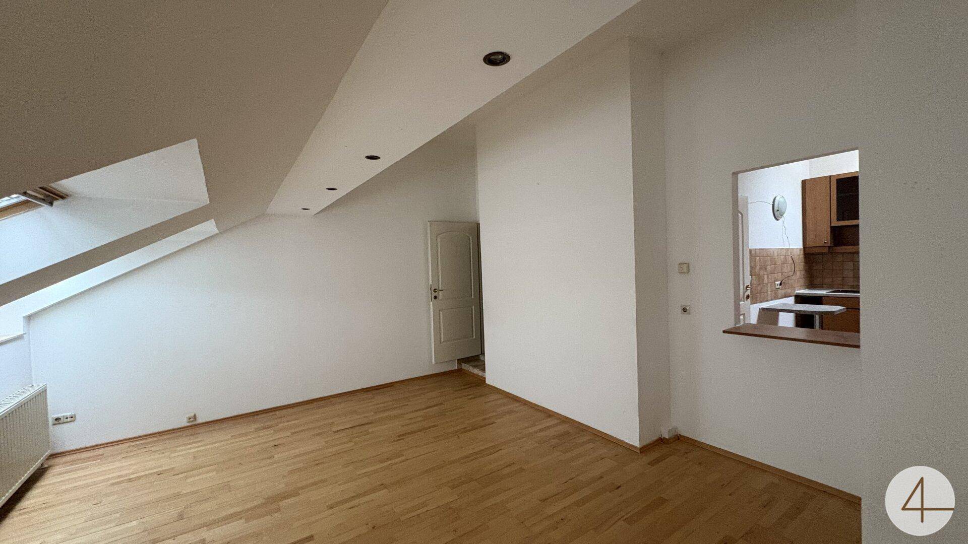 Dachgeschoss-Wohnzimmer mit Holzboden, Dachfenstern und Zugang zur Küche durch eine praktische Öffnung.