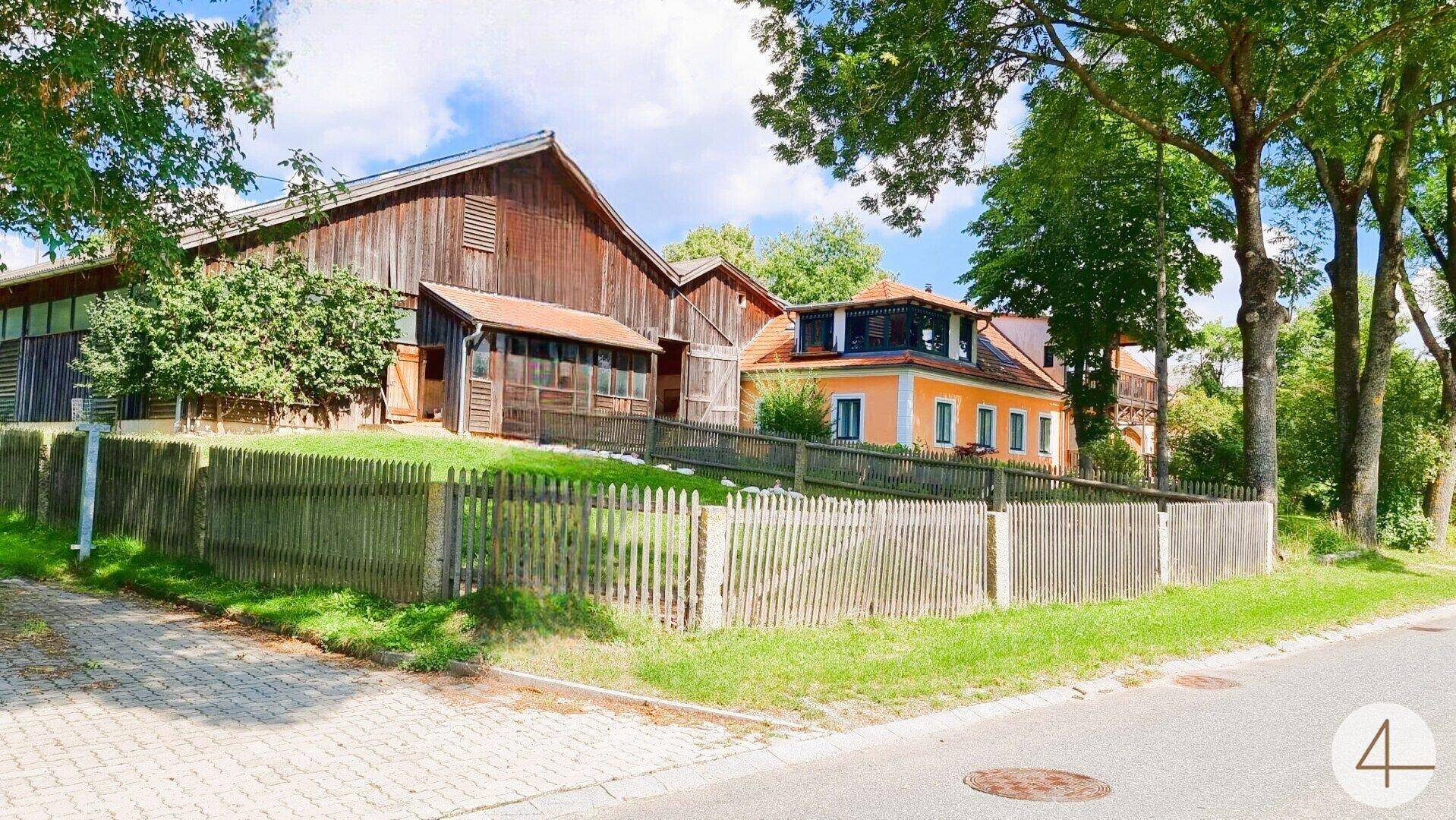 Anwesen mit traditionellen Gebäuden und Holzzaun entlang einer ruhigen Straße, umgeben von Bäumen.
