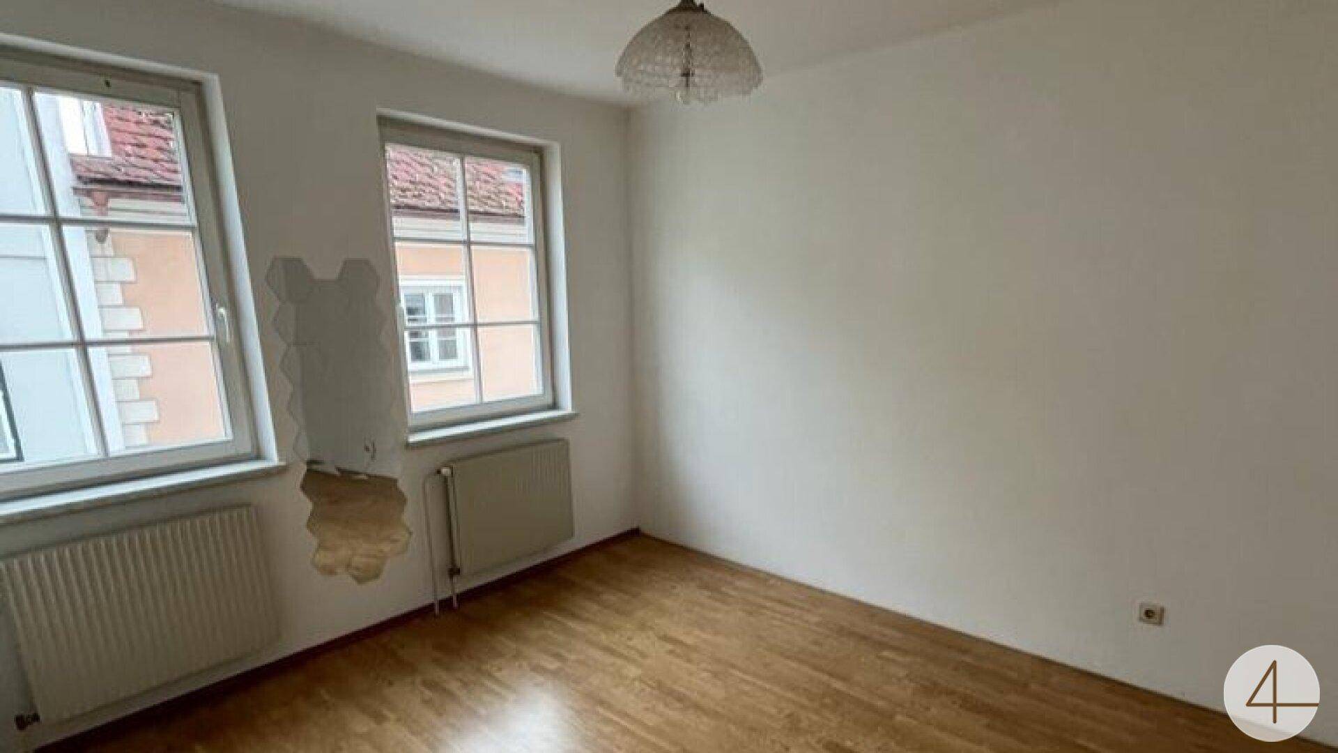 Leeres Zimmer mit Holzboden, zwei Fenstern und Blick auf gegenüberliegende Gebäude.