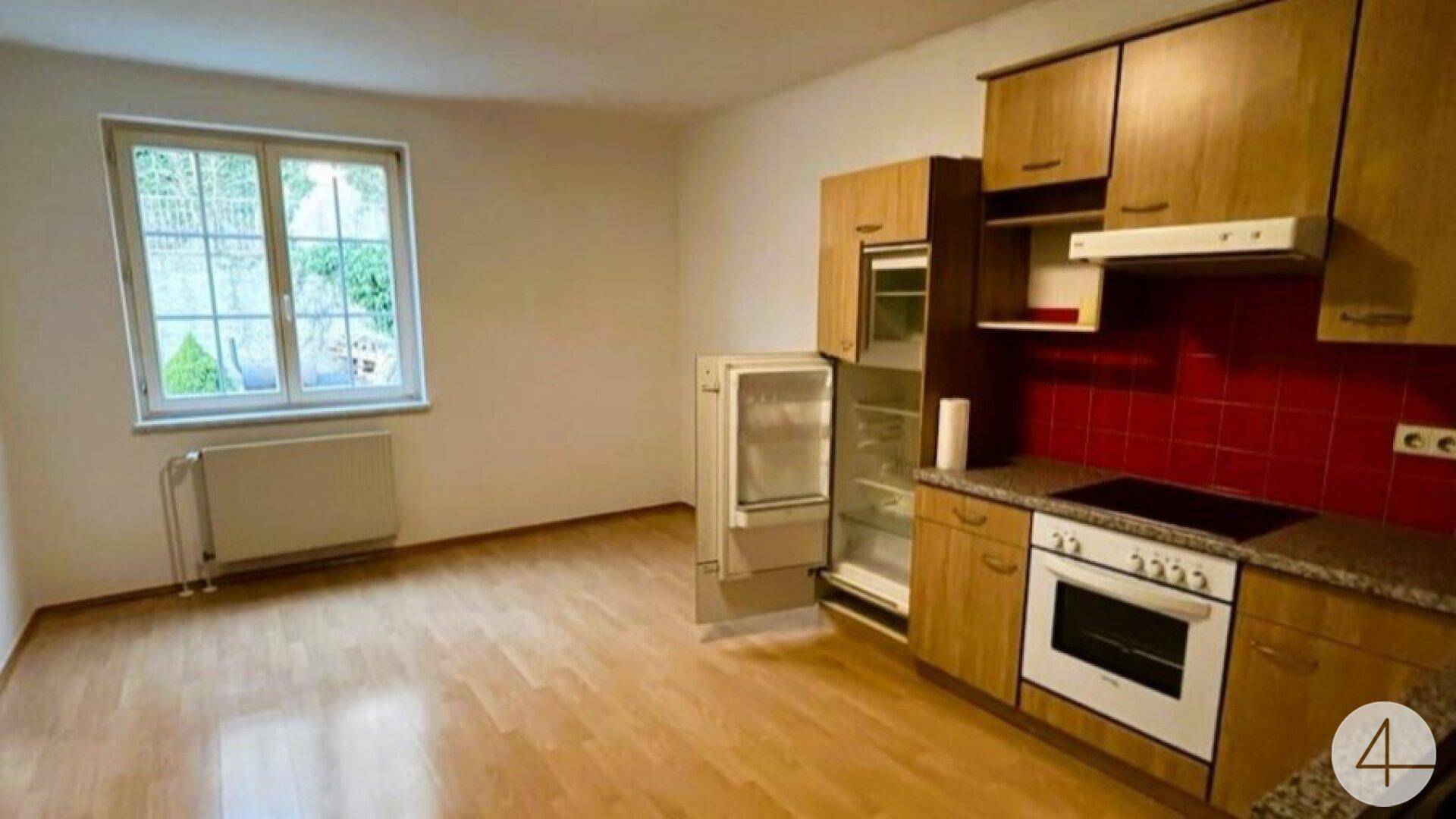 Einfache Küche mit Holzschränken, rotem Fliesenspiegel und Blick ins Grüne.