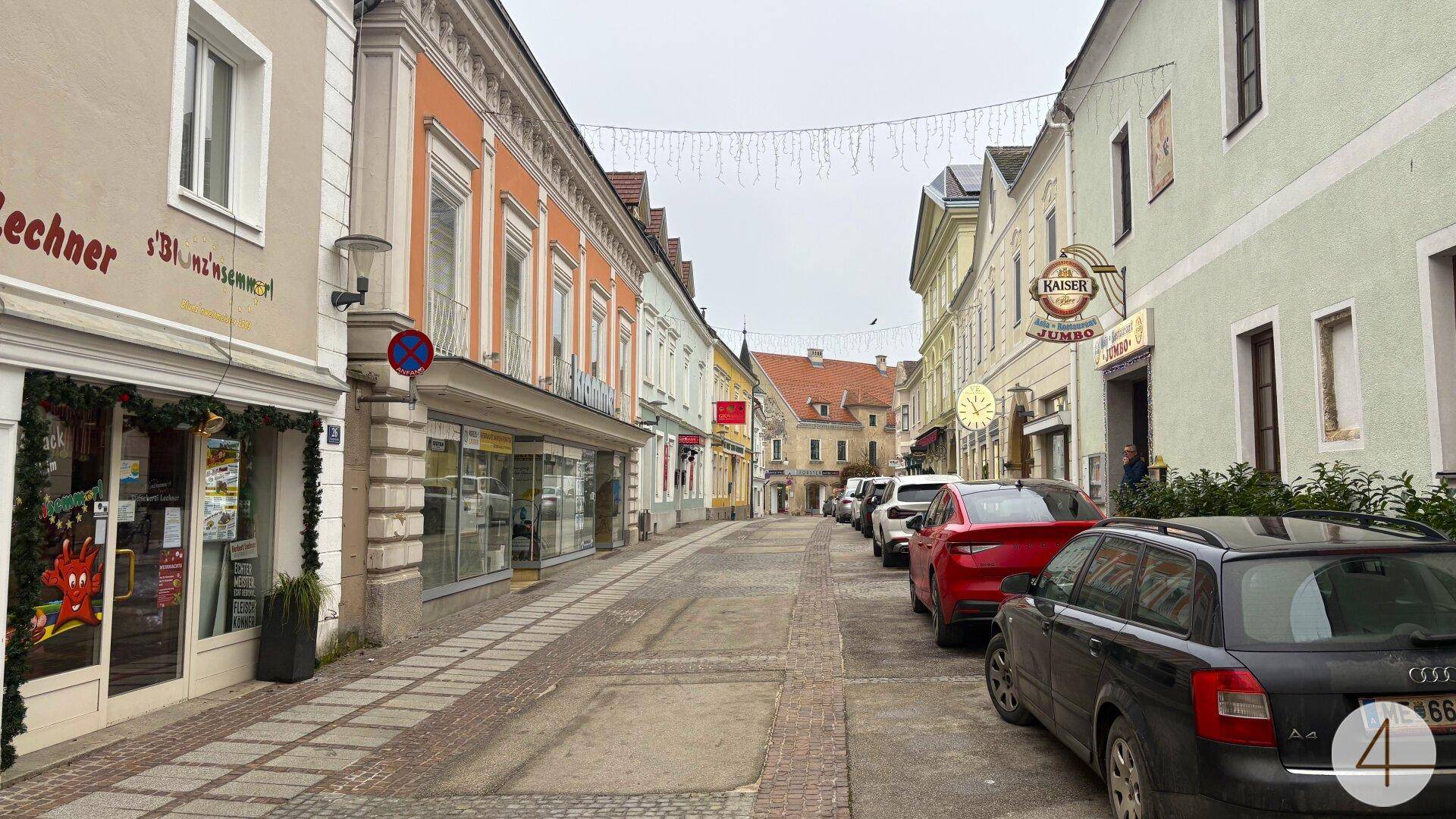Beleuchtete Einkaufsstraße mit parkenden Autos und Geschäften in historischen Gebäuden.