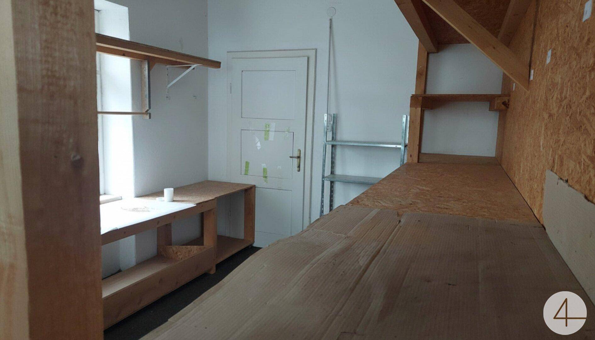 Dachbodenraum mit sichtbaren Holzbalken, Regalen und einem Fenster für Tageslicht.