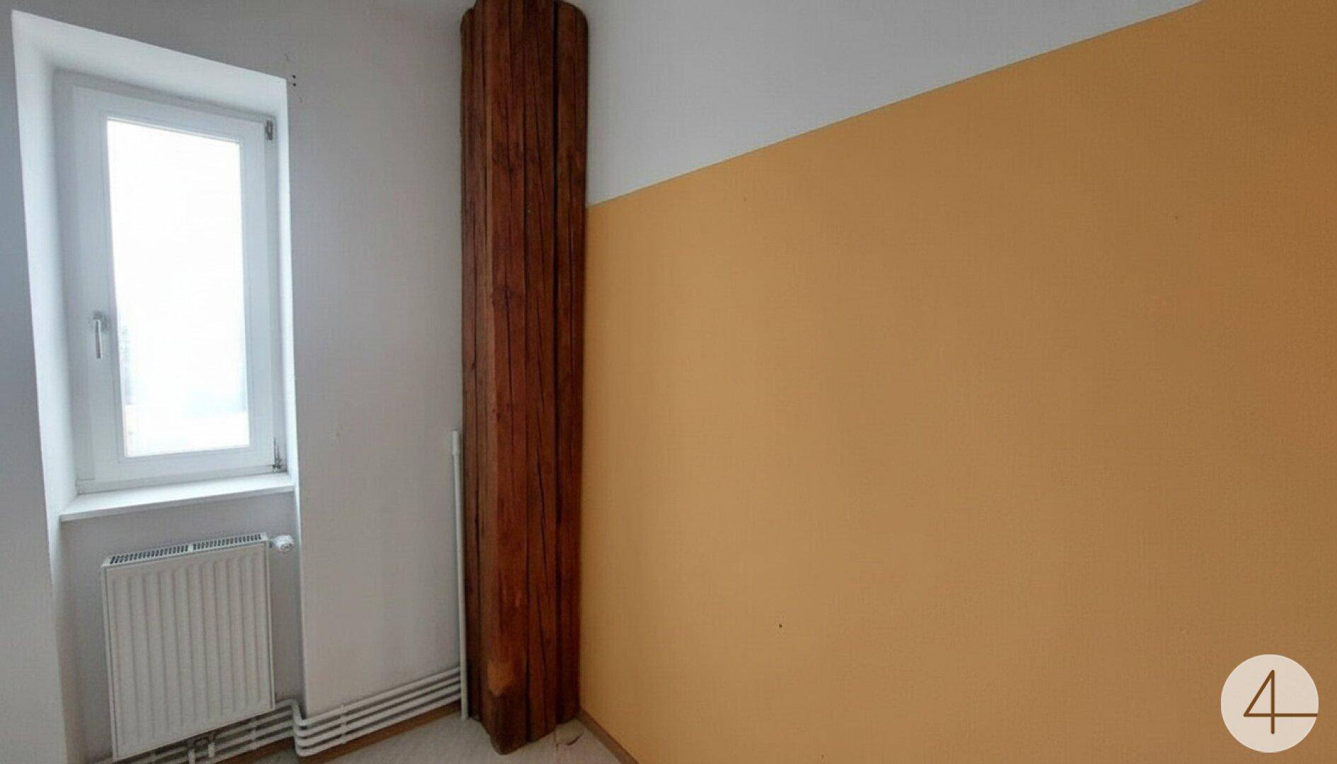 Kleiner Raum mit Fenster und einer auffälligen Holzverkleidung an der Wand, gelbe Wandfarbe.