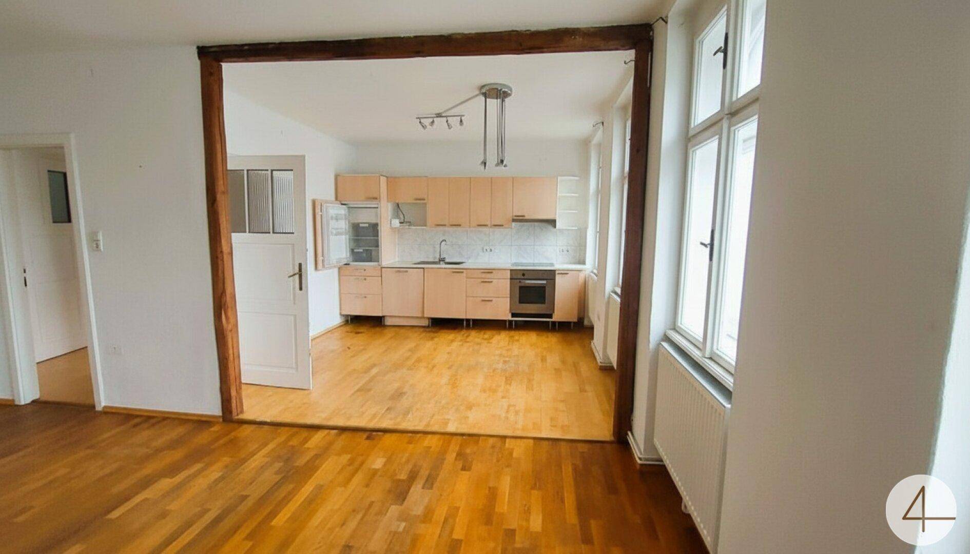 Heller Wohnbereich mit Holzboden und offener Verbindung zu einer Einbauküche mit hellen Schränken.