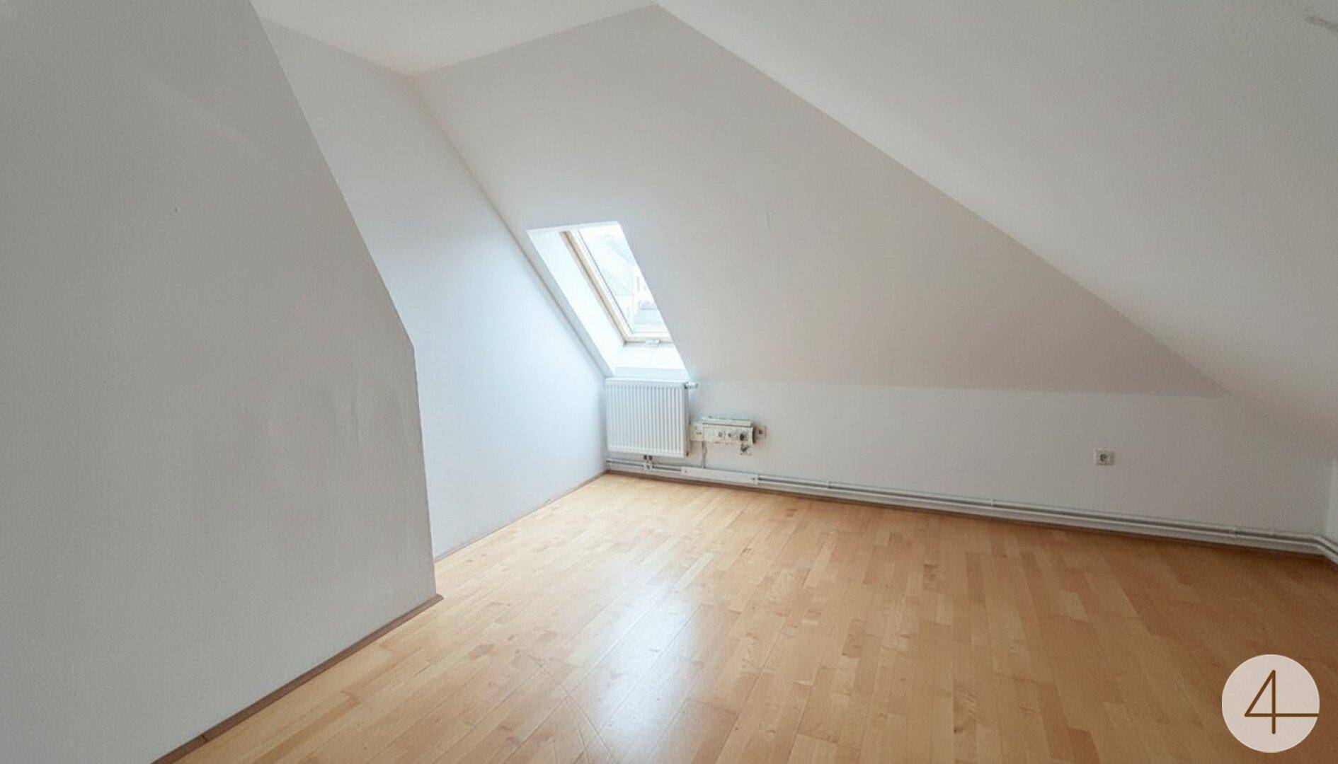 Dachzimmer mit schrägen Wänden, Laminatboden und einem Dachfenster, das viel Tageslicht hereinlässt.