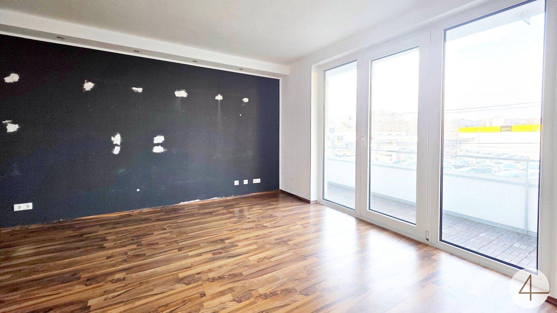 Großes Wohnzimmer mit Laminatboden und breiter Fensterfront, die Zugang zum Balkon ermöglicht.