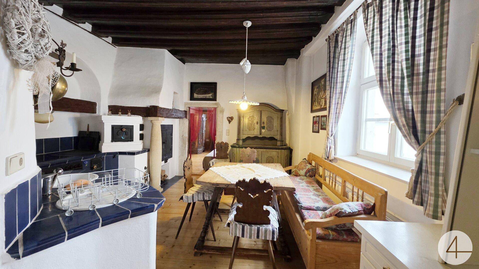 Rustikale Wohnküche mit traditionellem Kachelofen, Holztisch und Fenster mit Vorhängen.