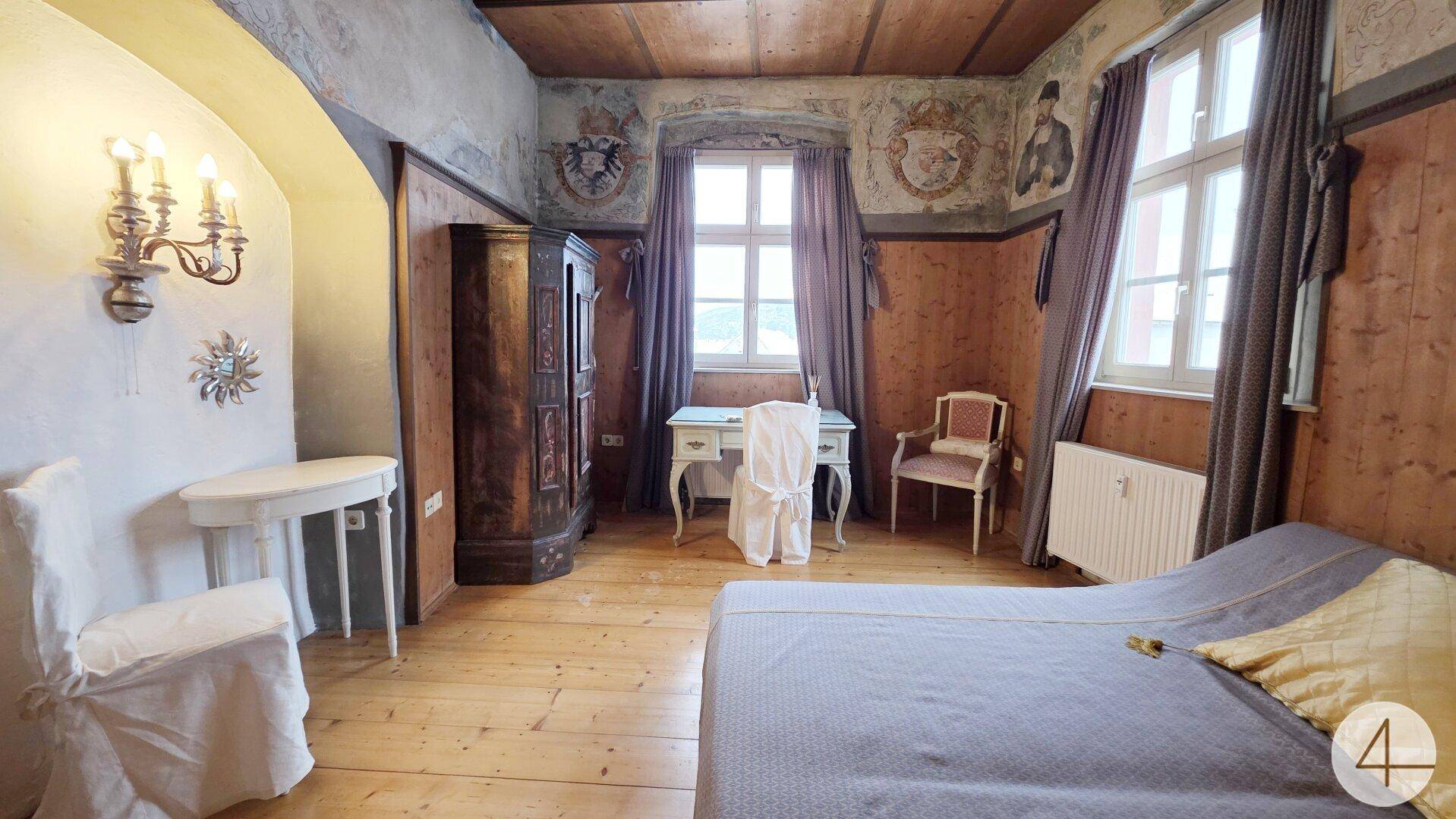 Historisches Schlafzimmer mit Holzwänden, bemalter Decke, Bett und Schreibtisch am Fenster.