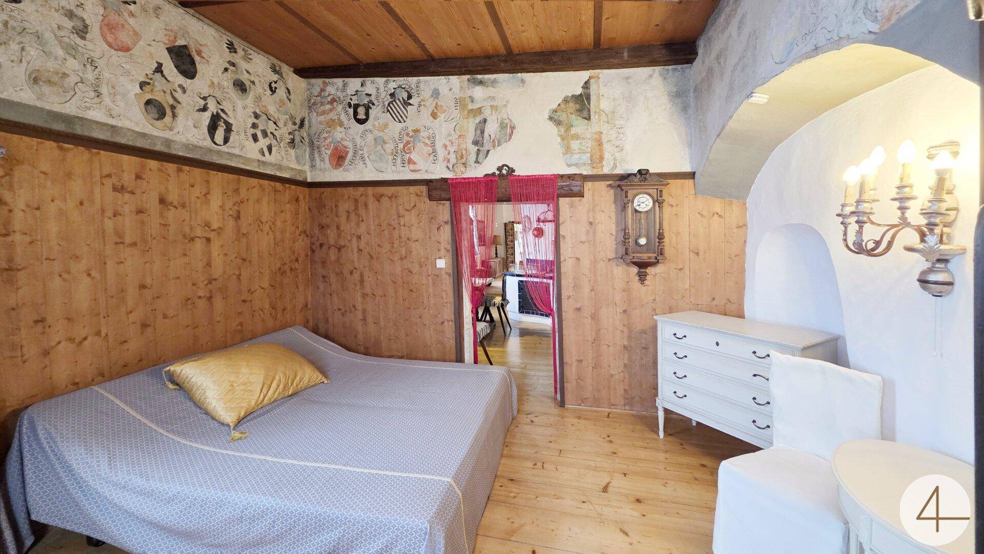 Gemütliches Schlafzimmer mit Holzwänden, bemalter Decke und einem Bett mit goldener Kissen.