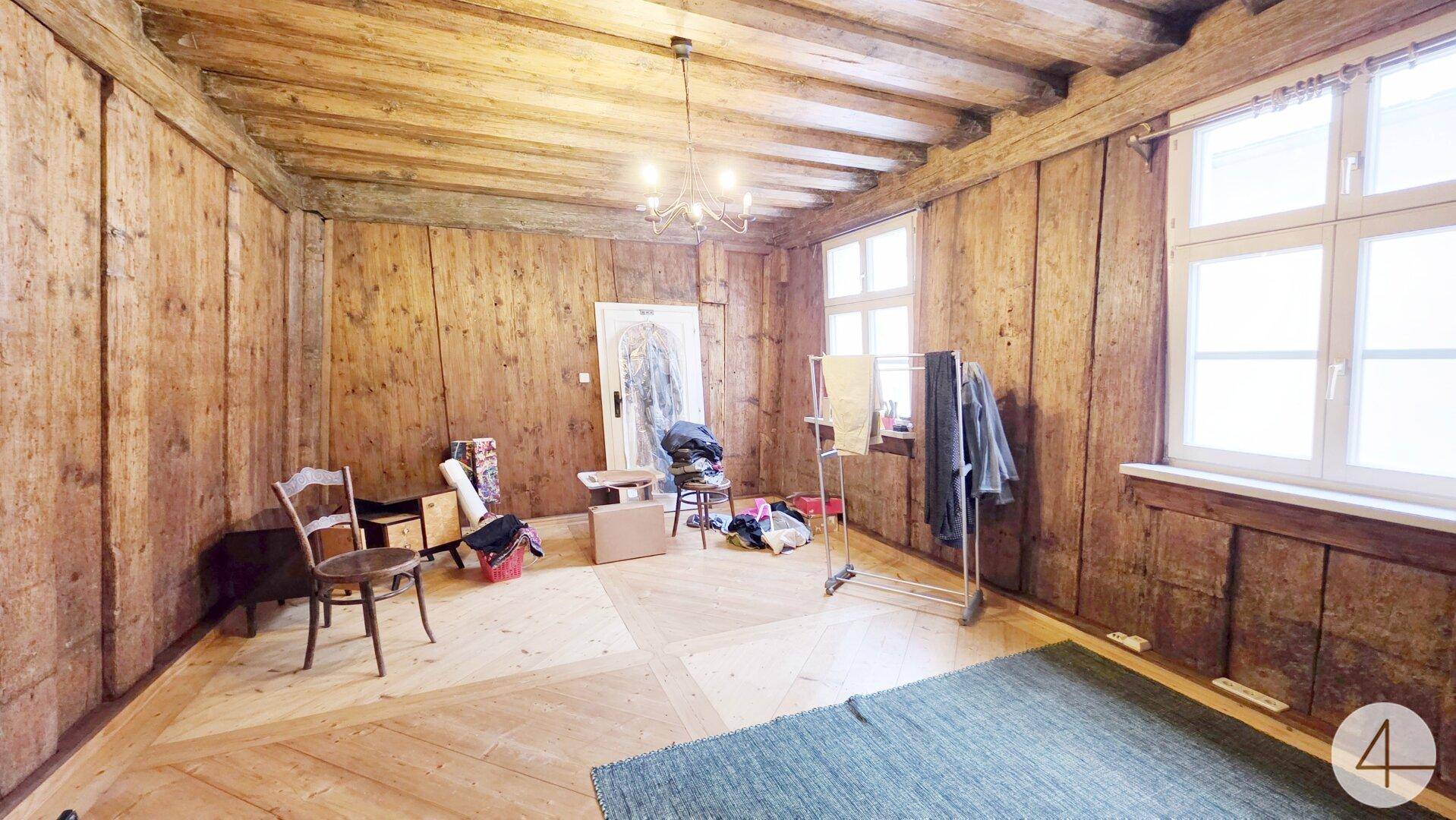 Zimmer mit rustikalen Holzvertäfelungen an Wänden und Decke sowie hellem Holzboden und Fenstern.