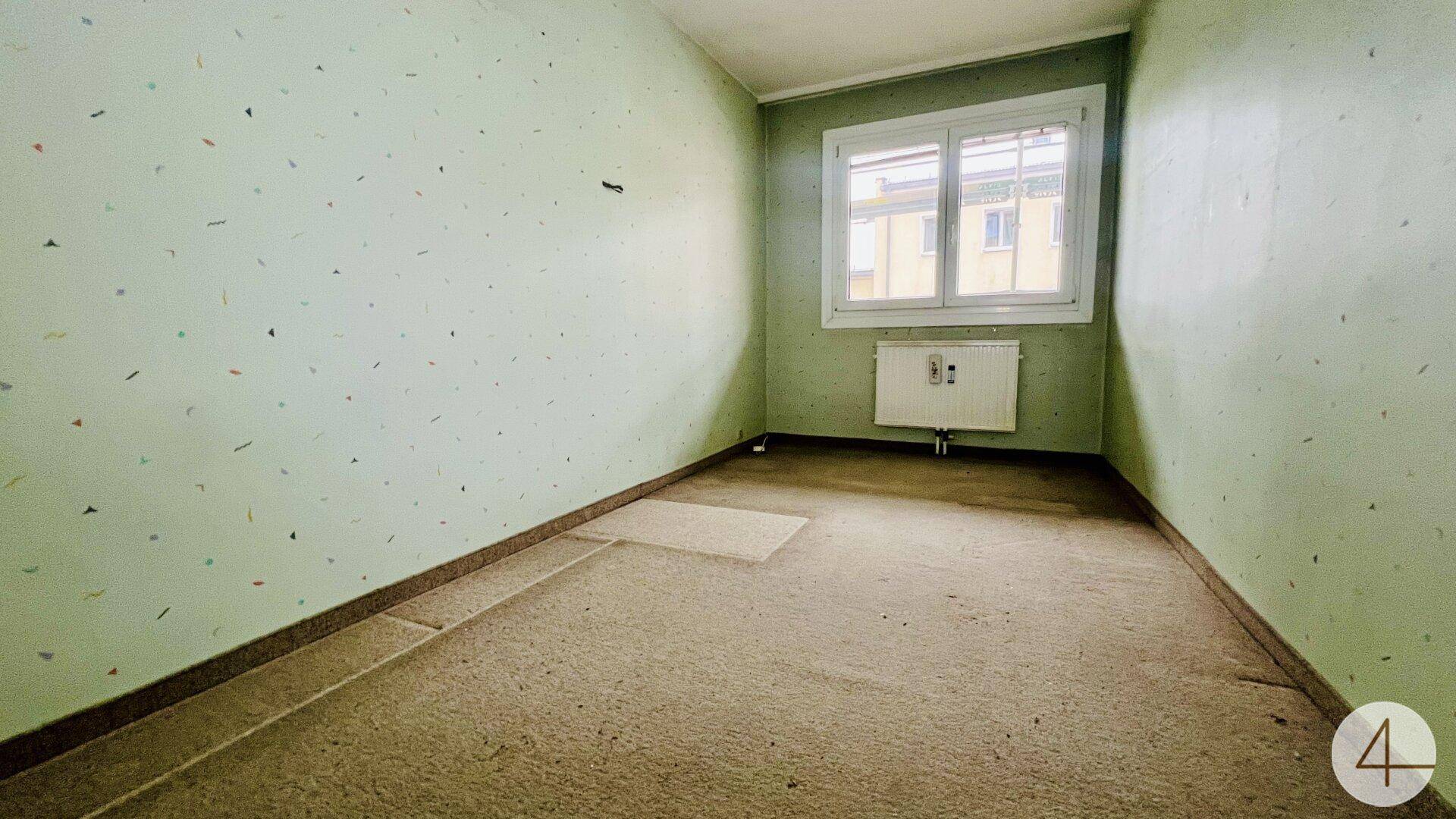 Leeres Zimmer mit grüner Mustertapete, Teppichboden und Heizkörper unter dem Fenster.