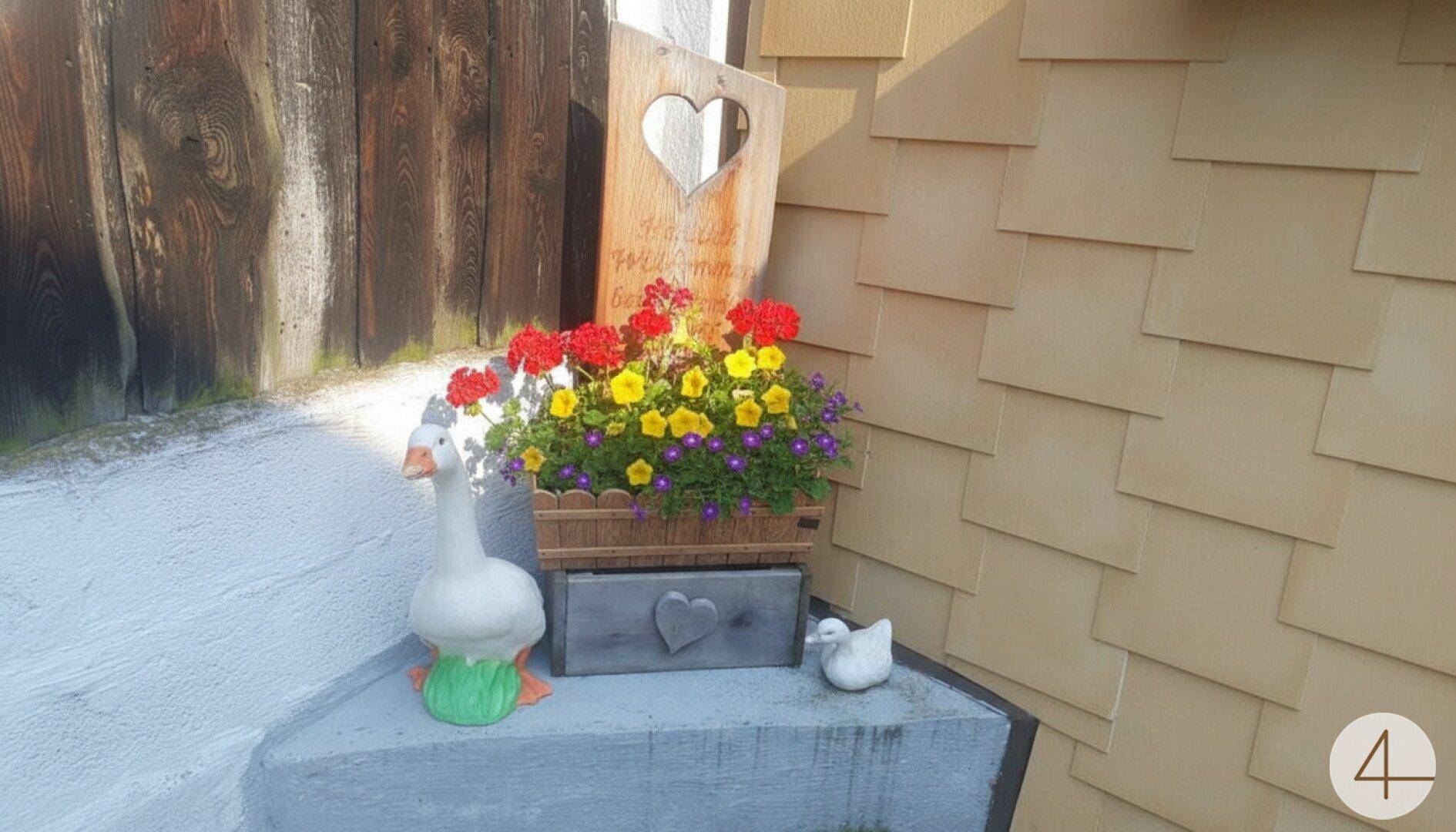 Liebevolle Außendekoration mit Blumen, einer Holztafel und Entenfiguren am Hauseingang.