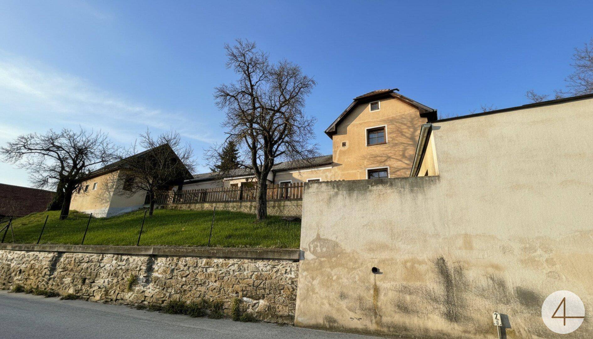Mehrere ältere Gebäude auf einem Hügel mit einer traditionellen Steinmauer im Vordergrund unter blauem Himmel.