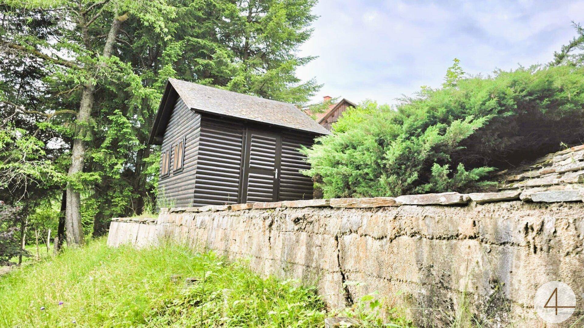 Kleines Holzhaus im Garten, umgeben von Bäumen und einer Steinmauer.