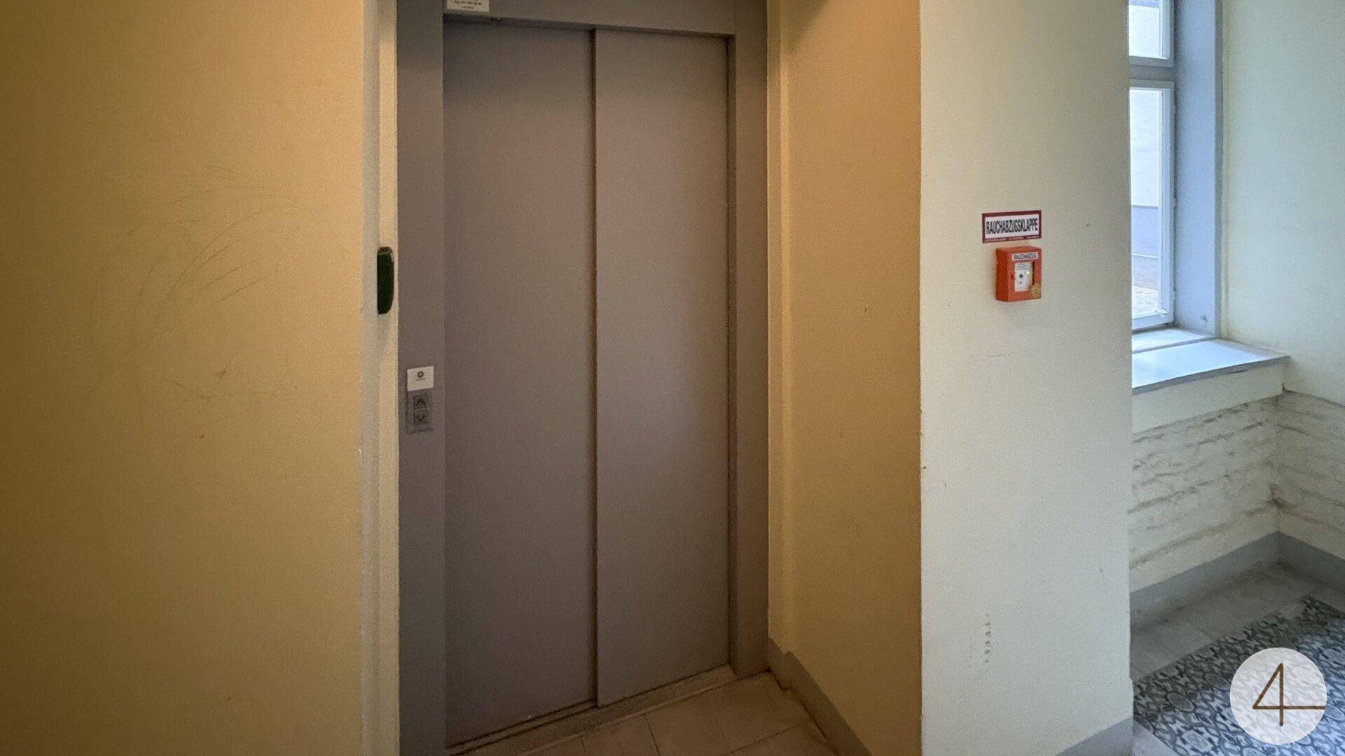 Heller Flur mit Aufzug, Fenster und Sicherheitseinrichtungen im Treppenhaus.